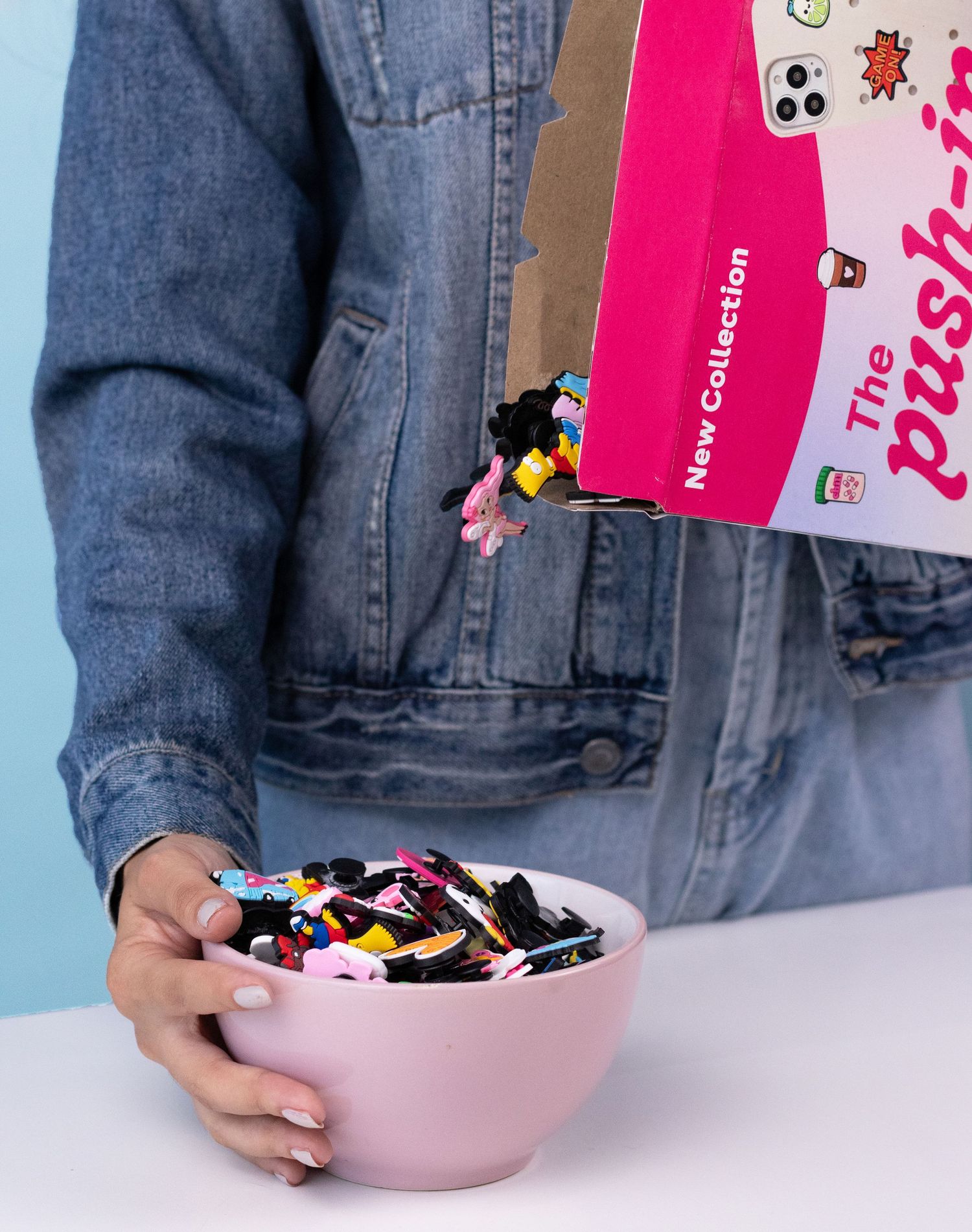 Person in denim jacket pouring colorful hair accessories from pink box into a pink bowl.