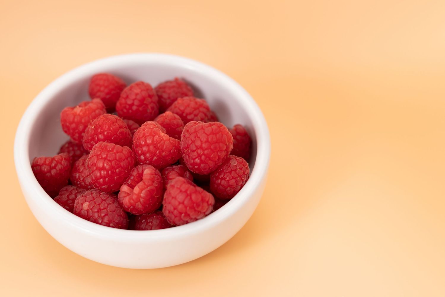 Fresh red raspberries in a white ceramic bowl on a soft orange background.