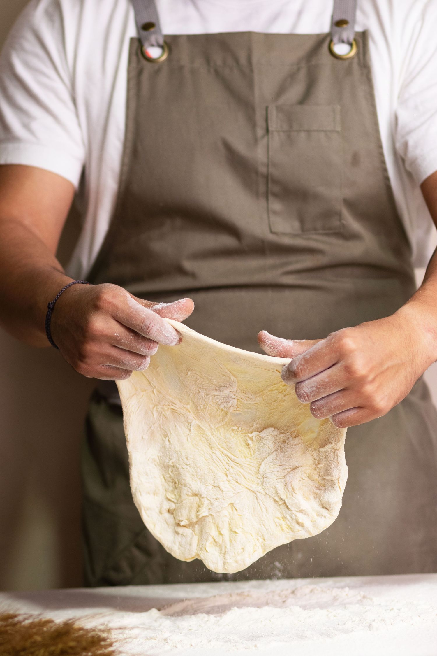 Baker in brown apron stretching and shaping fresh pizza dough on floured surface.