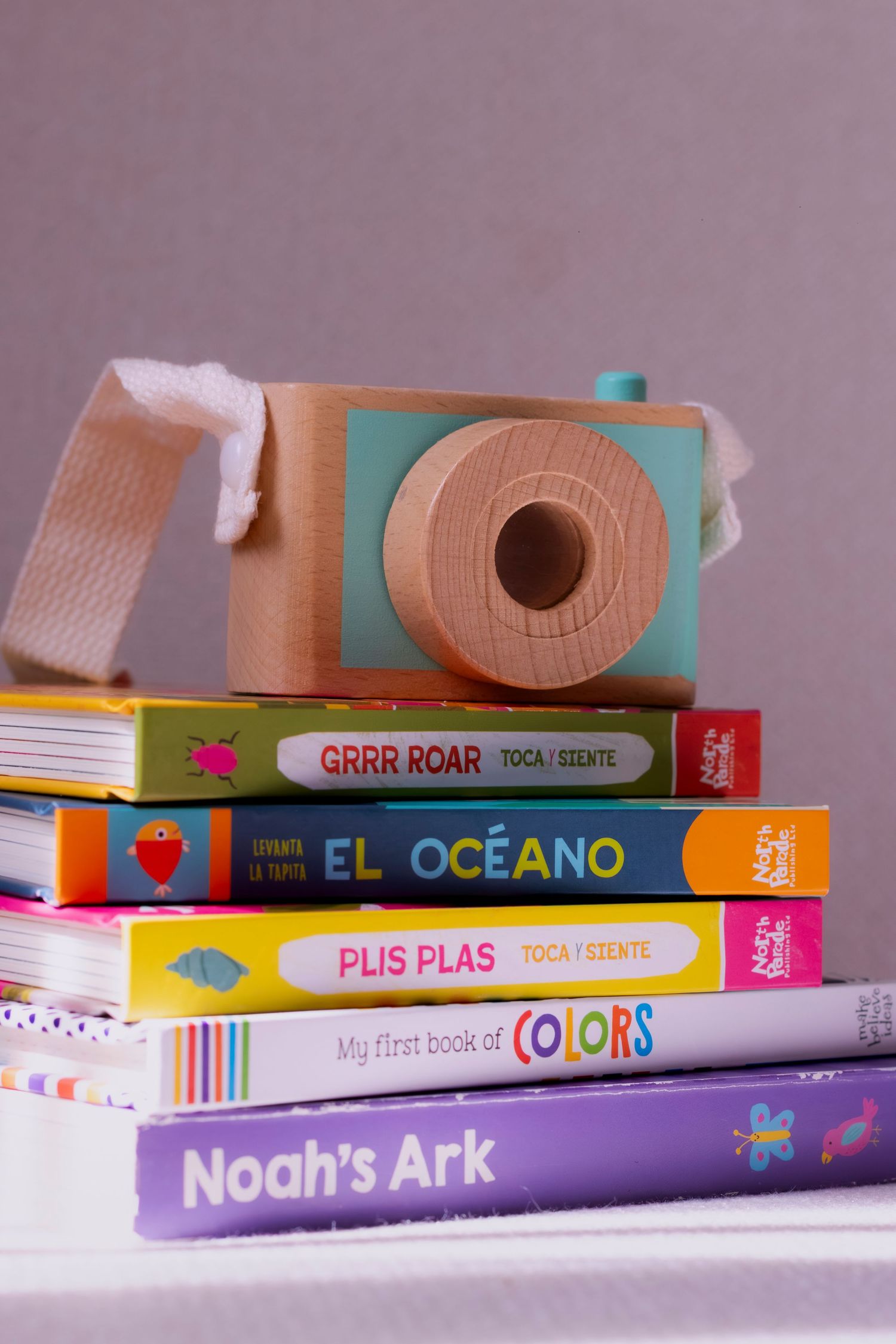 Stack of colorful children's books including Noah's Ark and El Oceano, topped with a wooden toy camera.