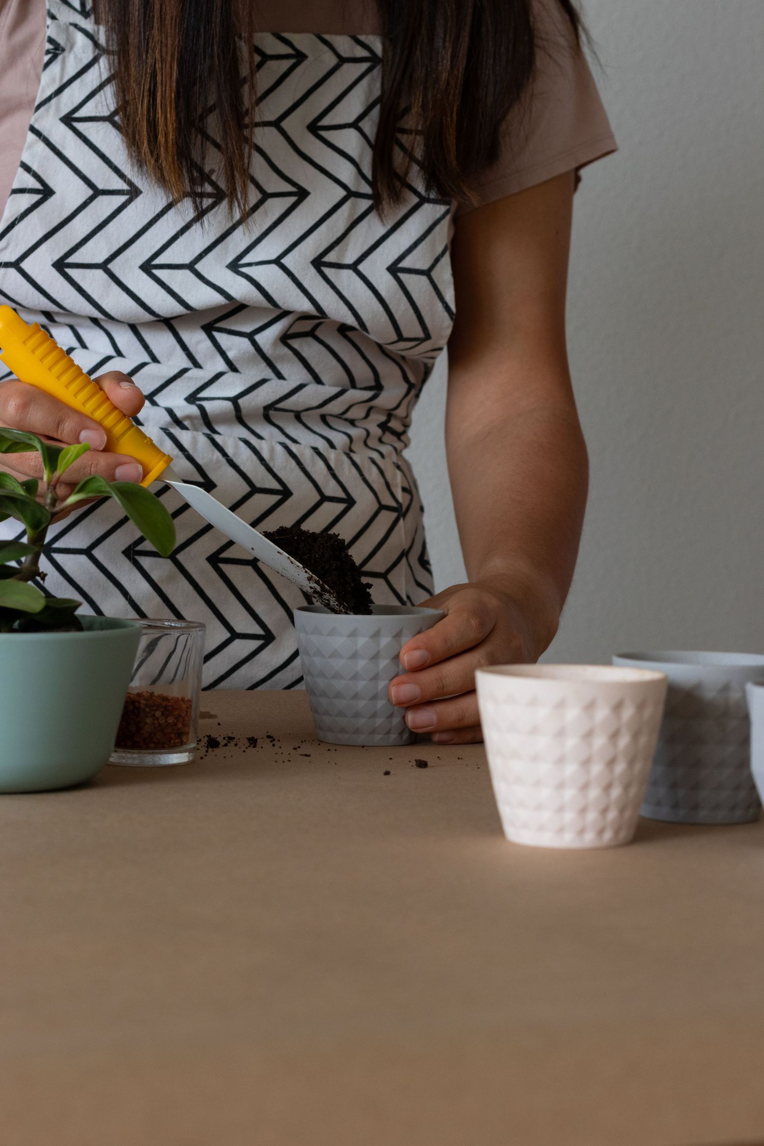 Woman in patterned apron repotting a plant into a gray textured pot on a wooden table.