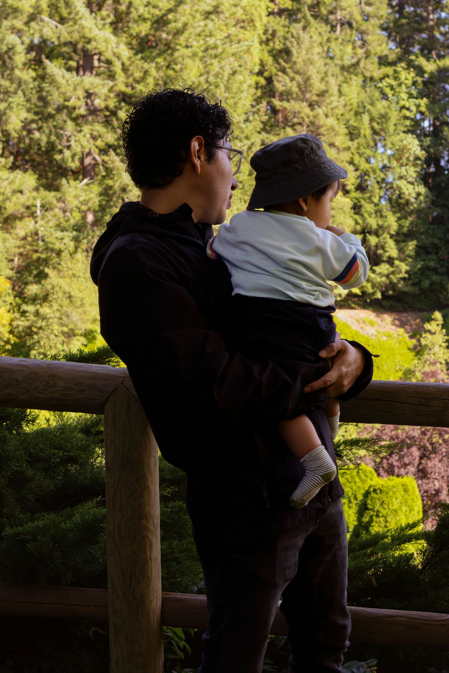Two people in dark clothing lean against a wooden railing in a lush outdoor setting with evergreen trees in background.