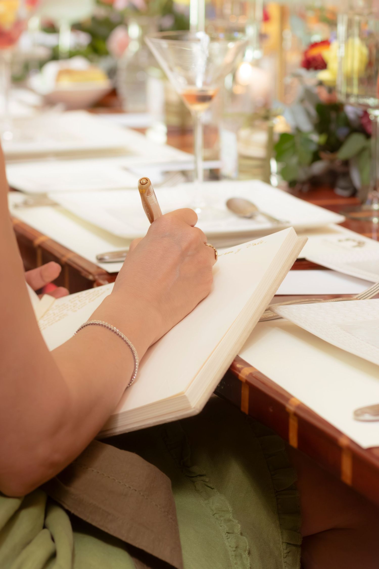 A woman writes in a notebook at an elegantly set dining table with wine glasses and floral centerpieces.