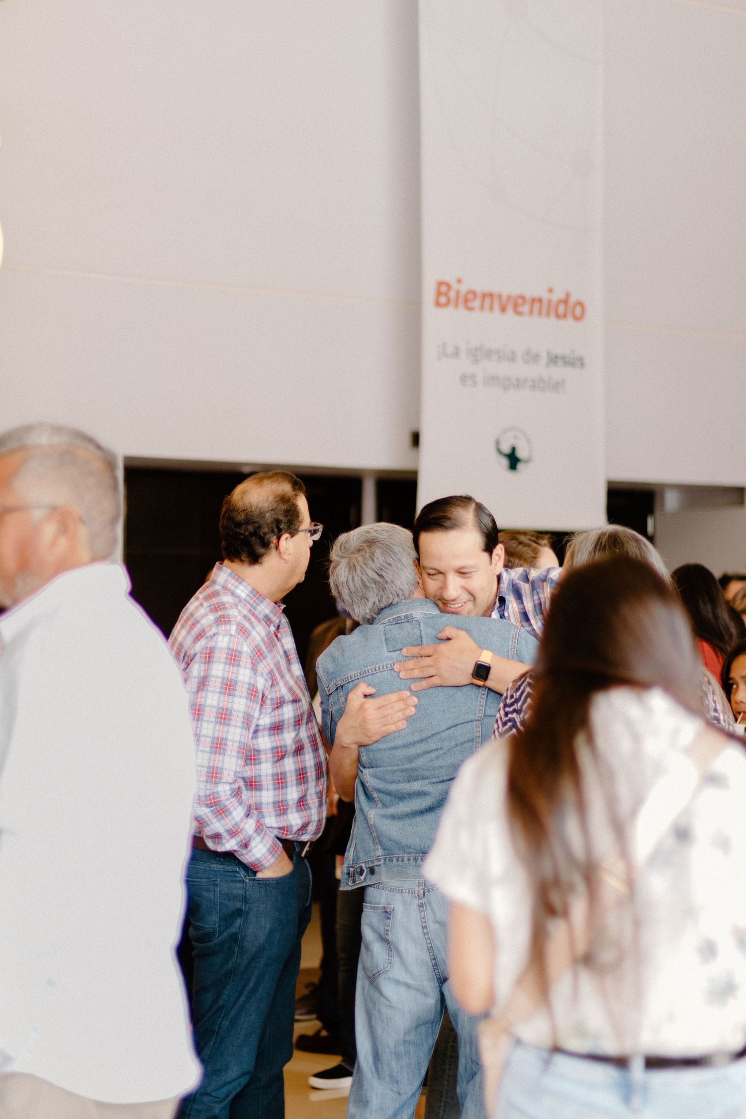 Men hugging warmly at a Spanish church welcome event with a Bienvenido sign in the background.