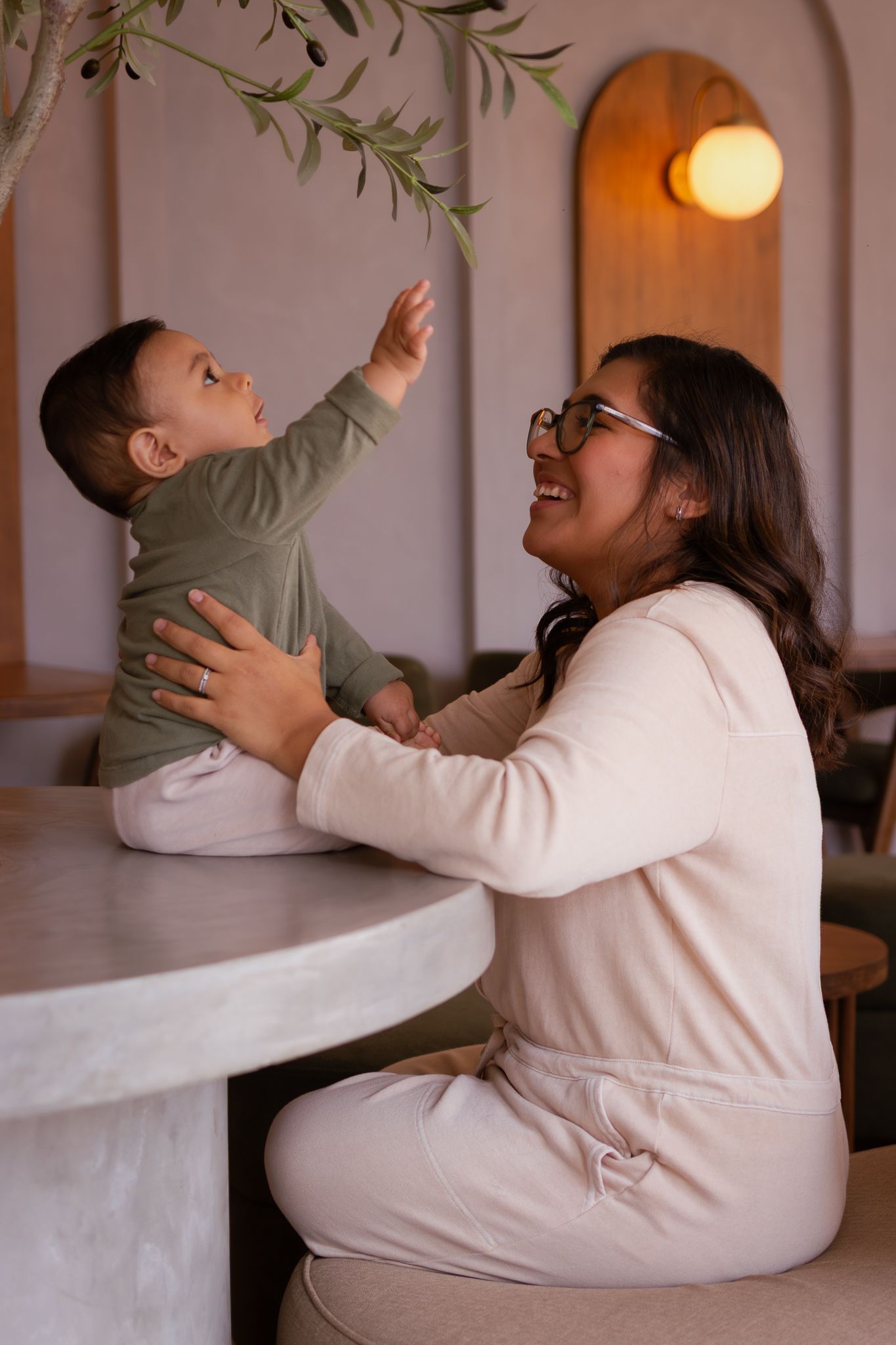 A smiling mother holds her baby reaching for olive tree branches at a cozy cafe table.