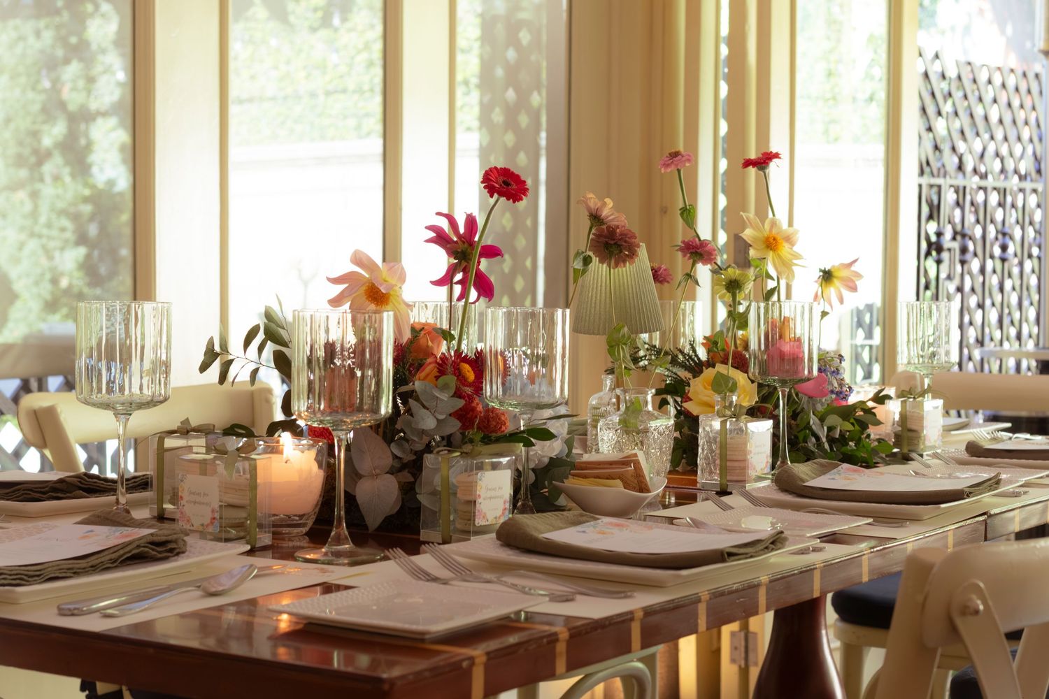Elegant dining table set with colorful floral centerpieces, candles, and fine glassware in a bright, airy room.