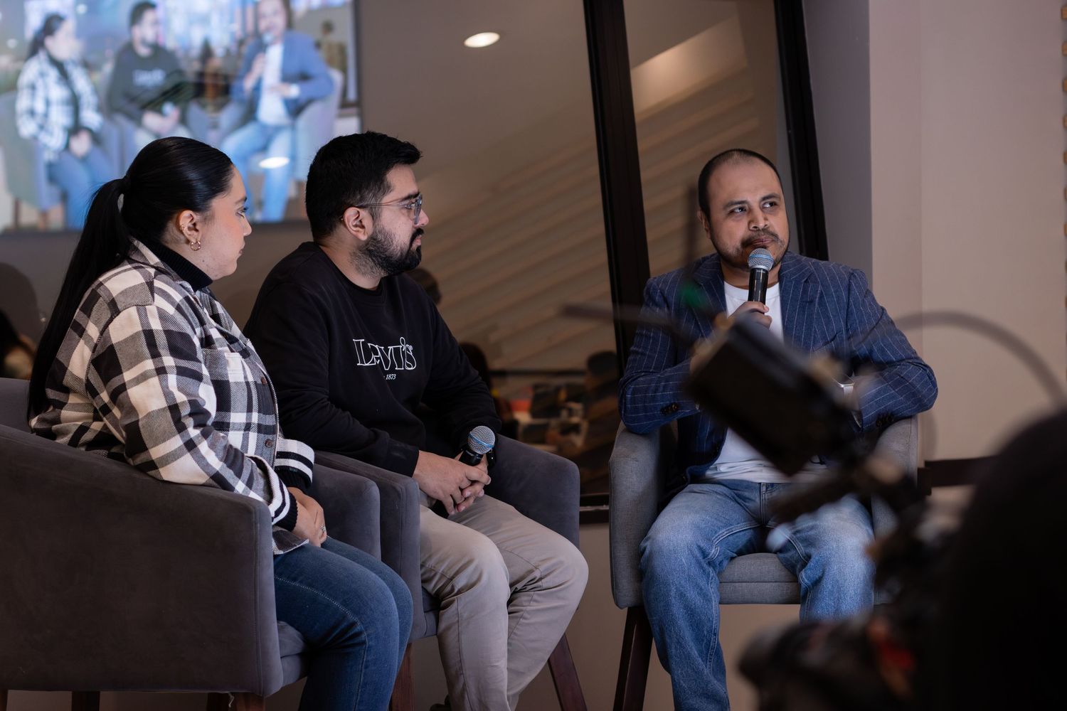 Three professionals engaged in a panel discussion, seated on chairs with a microphone, in a modern conference setting.