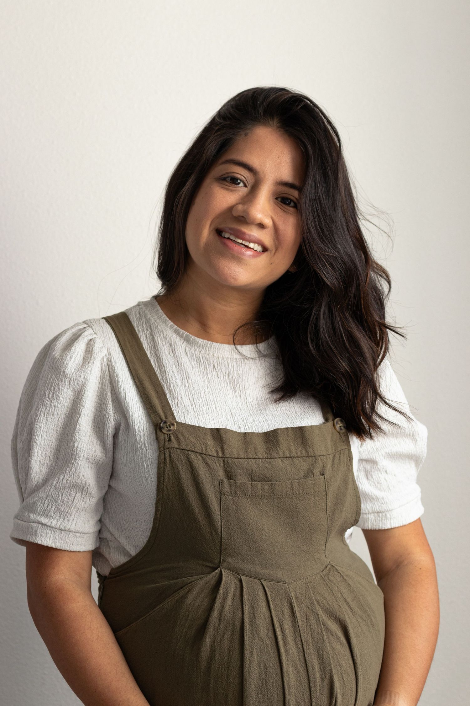 Smiling woman with dark hair wearing white blouse and olive green overalls against light background.