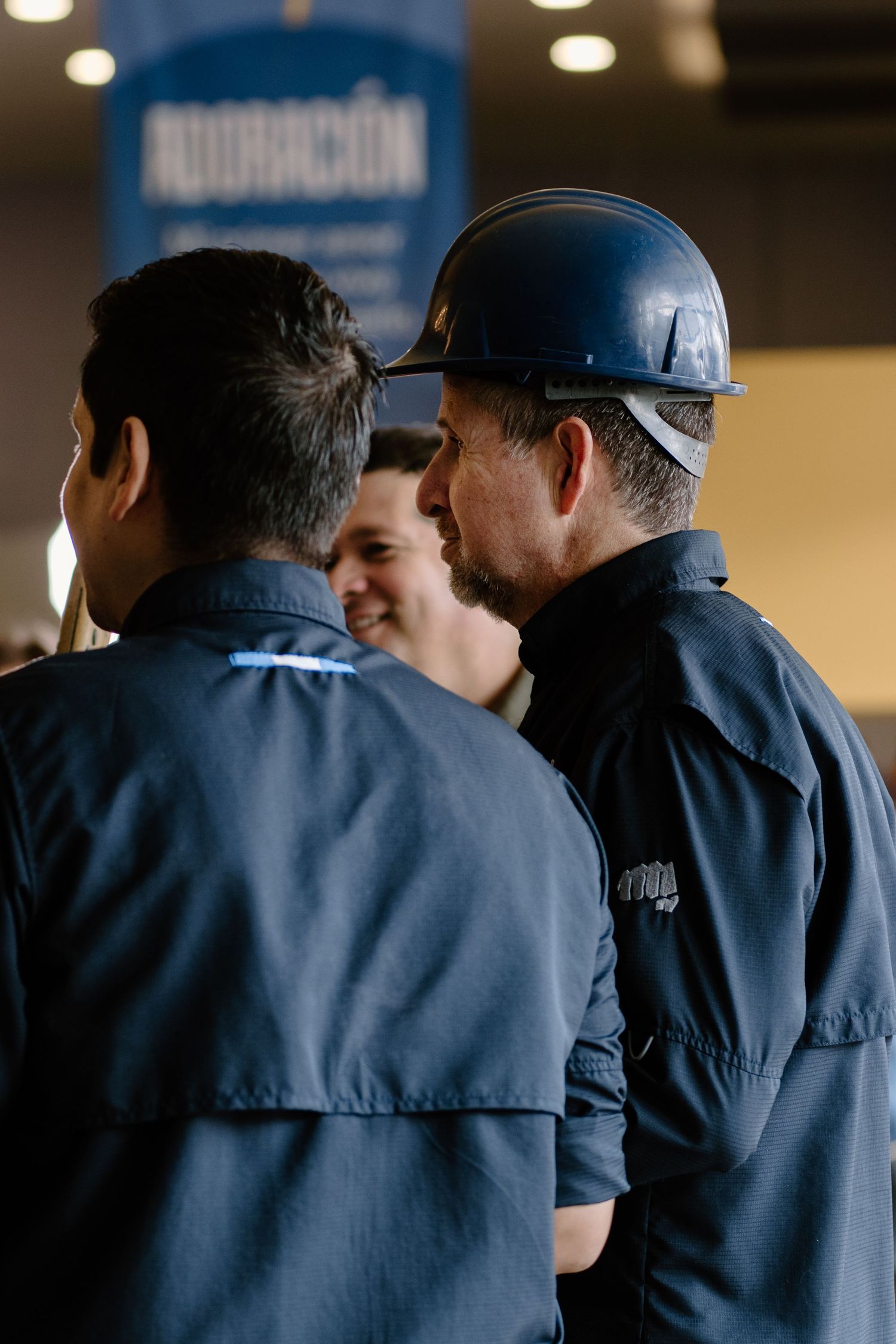 Workers in blue uniforms and hard hat conversing at an indoor industrial event with a banner in the background.