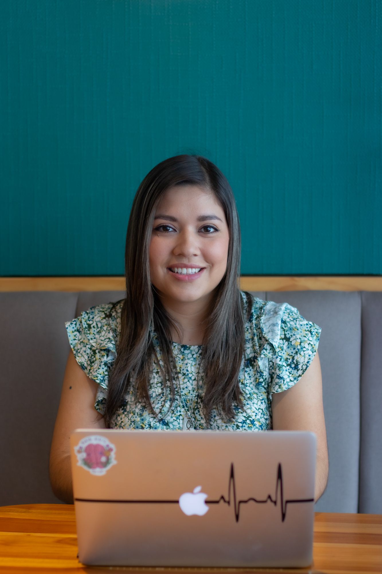 Smiling young woman sitting at wooden table with Apple laptop featuring heartbeat sticker, teal wall background.