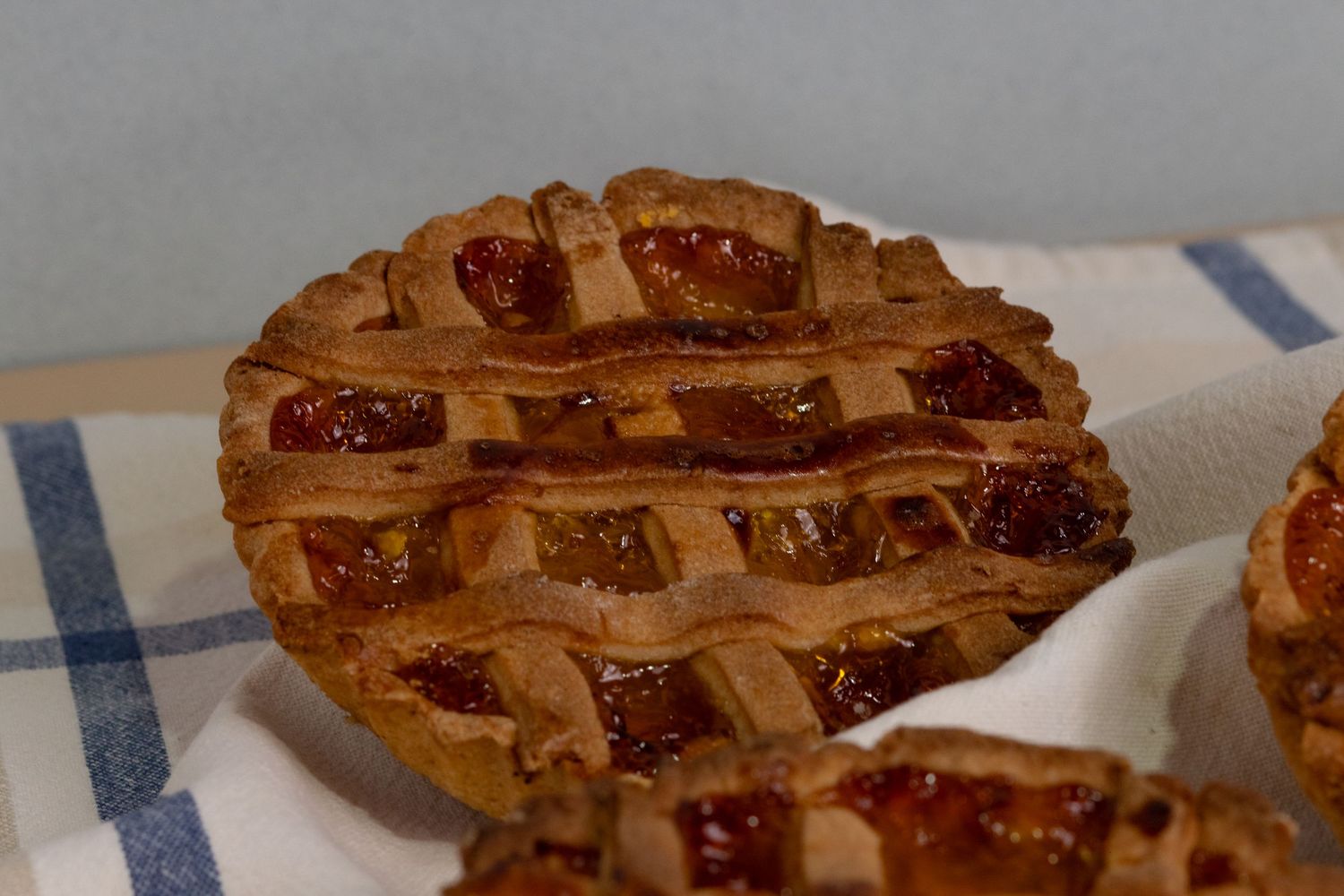 A golden-brown lattice-top mini fruit pie with glossy jam filling, resting on a blue and white checkered cloth.