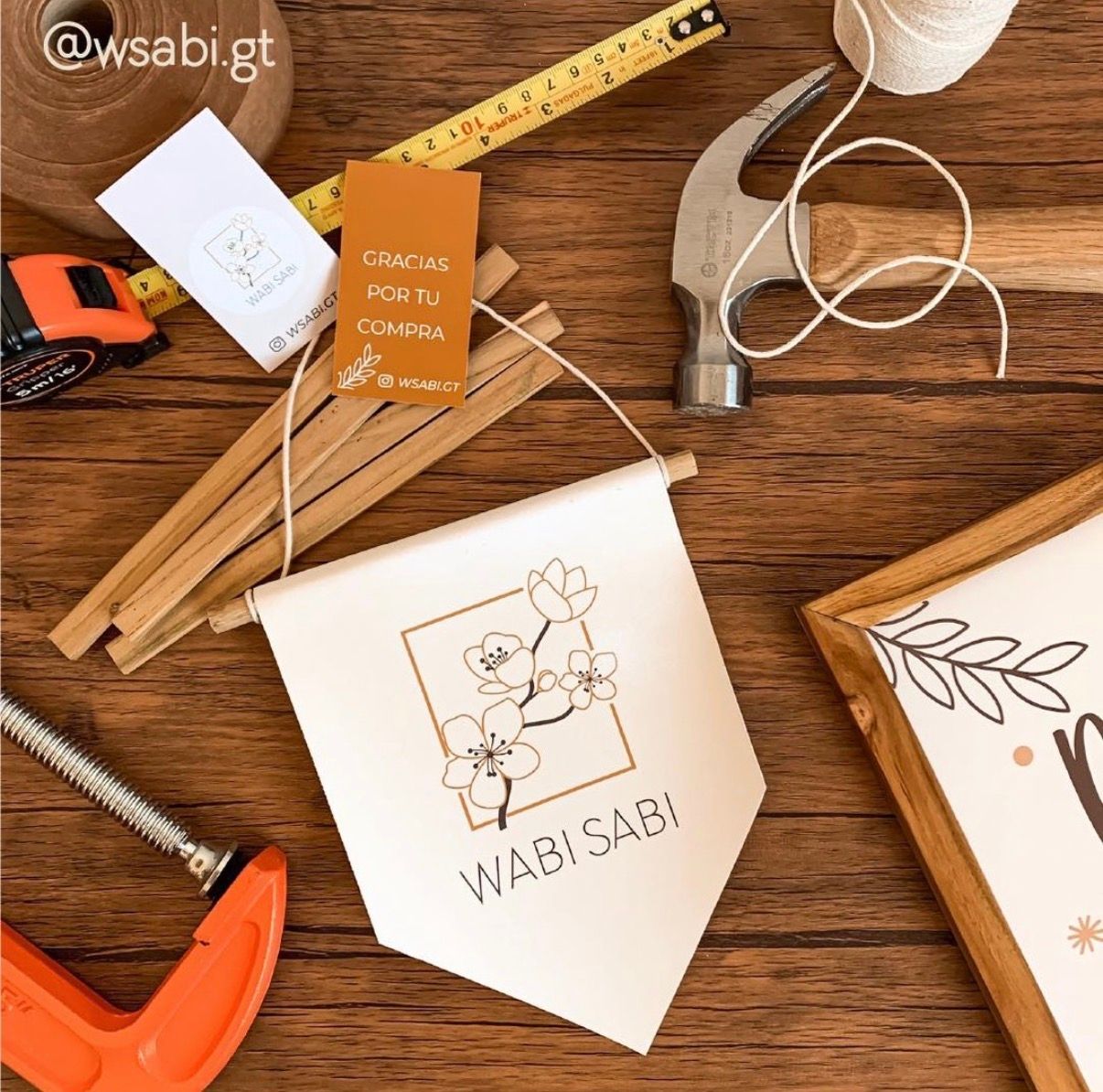 Wabi Sabi branded pennant flag displayed on wooden table with tools including hammer, tape measure, and clamps.
