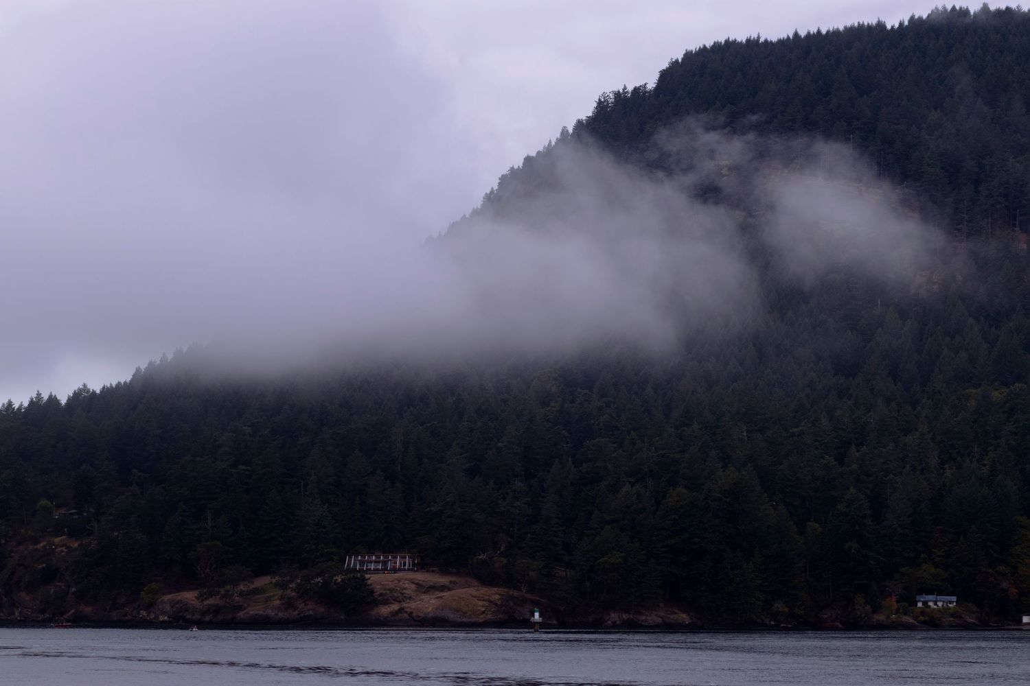 Low clouds drift across forested mountainside above calm waters with small buildings visible along shoreline.