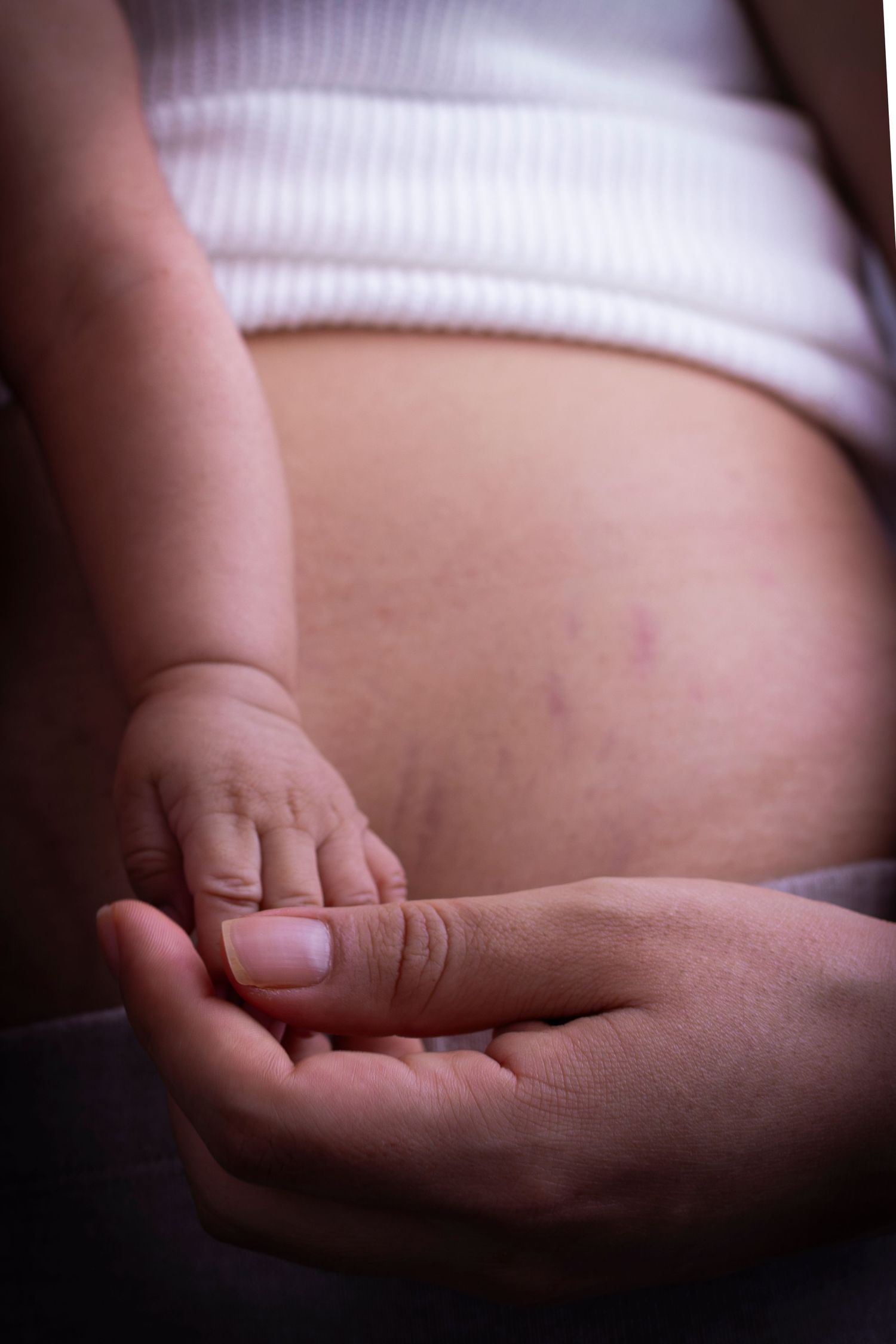 Gentle hands cradling a tiny baby hand against a pregnant belly in soft lighting.