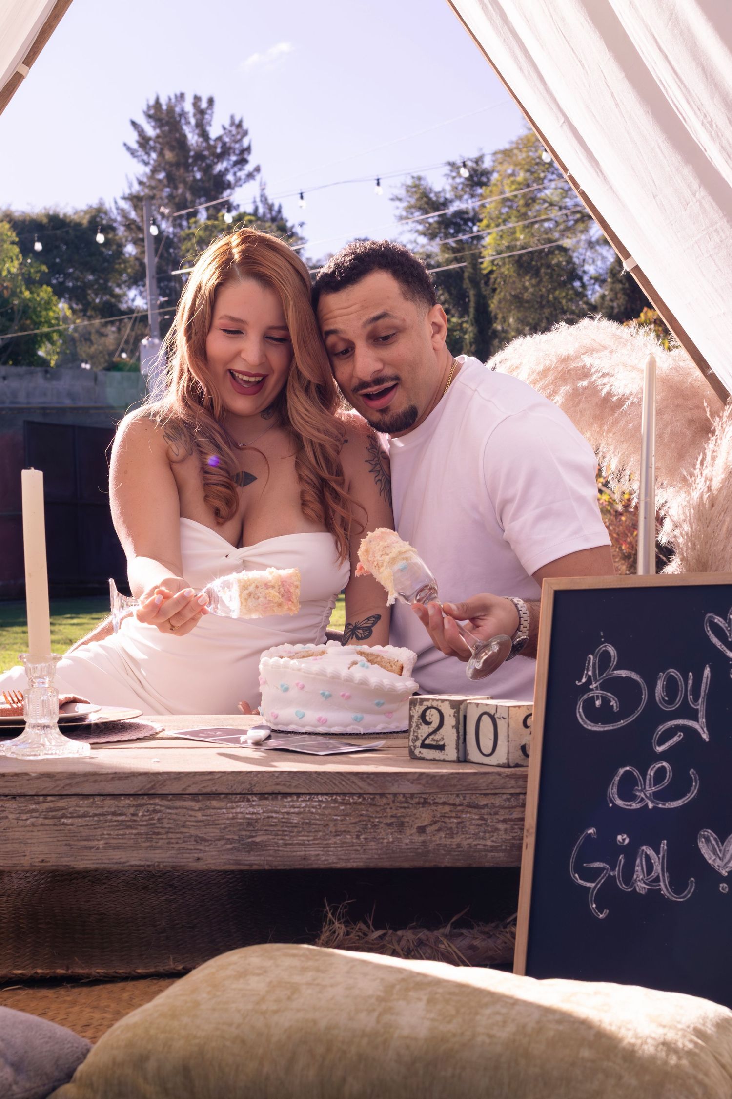 Couple cutting a white cake at a gender reveal party with a Boy or Girl chalkboard sign outdoors.