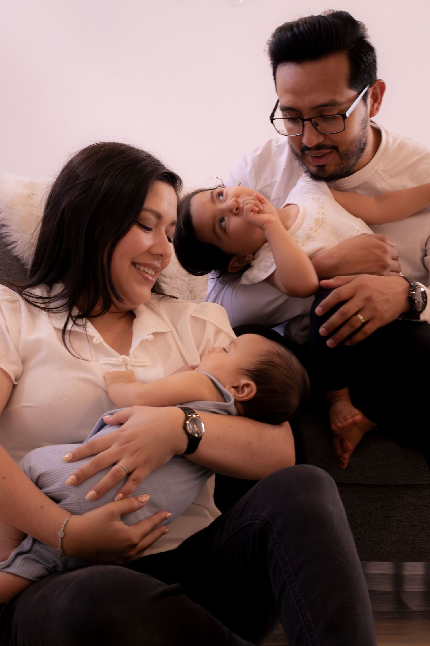 Happy Asian family relaxing on sofa, mother holding baby while father smiles at young child lying across their laps.