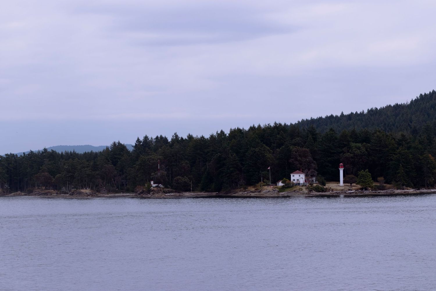 A white lighthouse stands on a forested coastline with calm waters and misty skies in the Pacific Northwest.