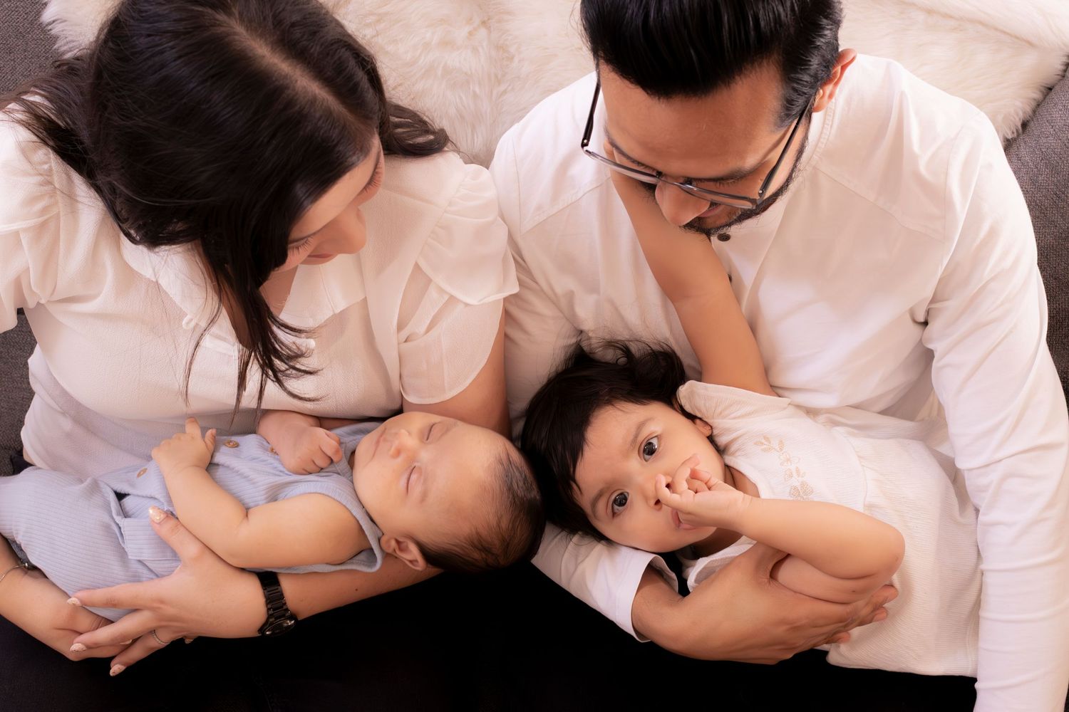 Asian family of four with parents in white shirts lovingly gazing at newborn baby and toddler child.