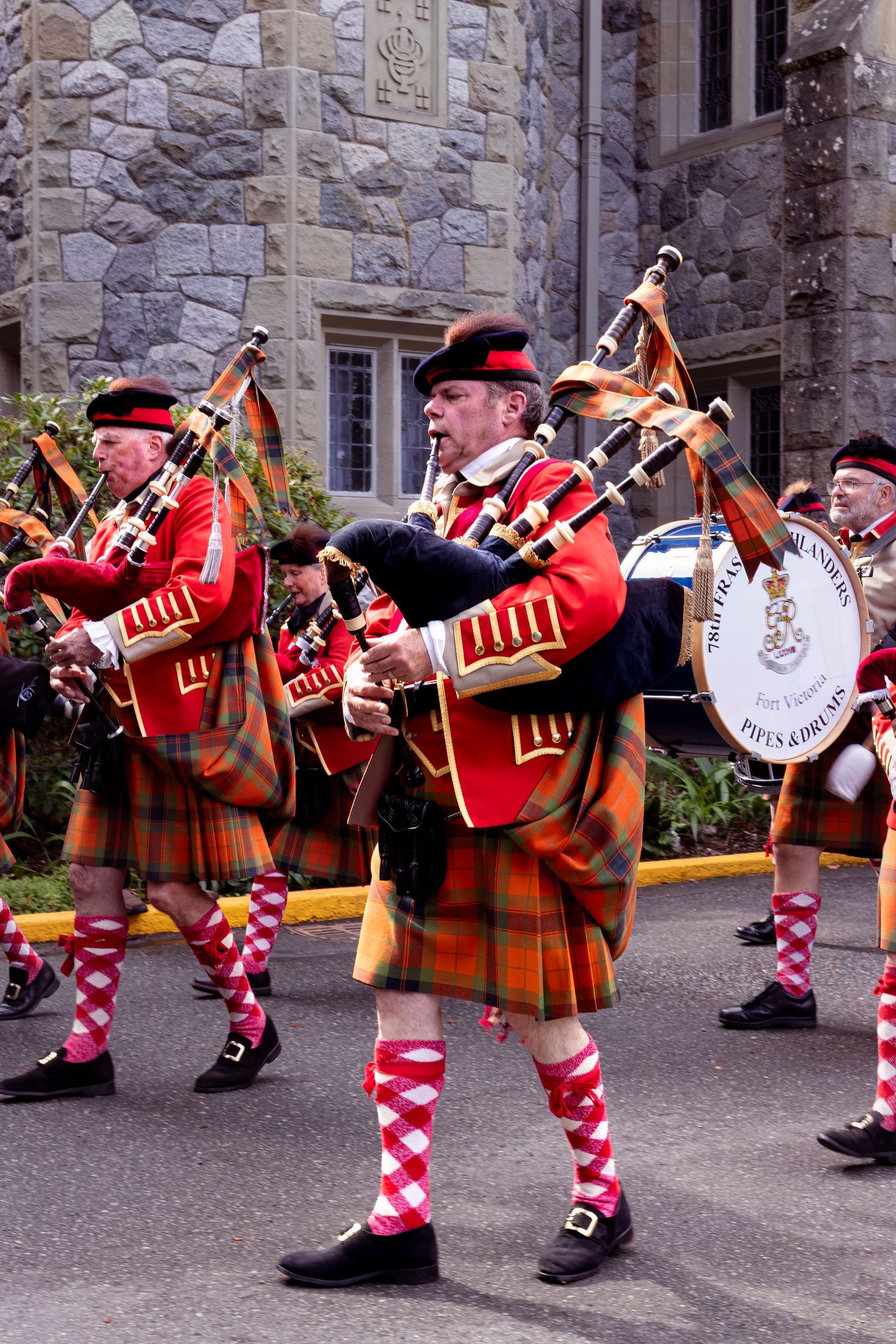 Scottish bagpipe band performs in traditional kilts and red jackets outside stone building.