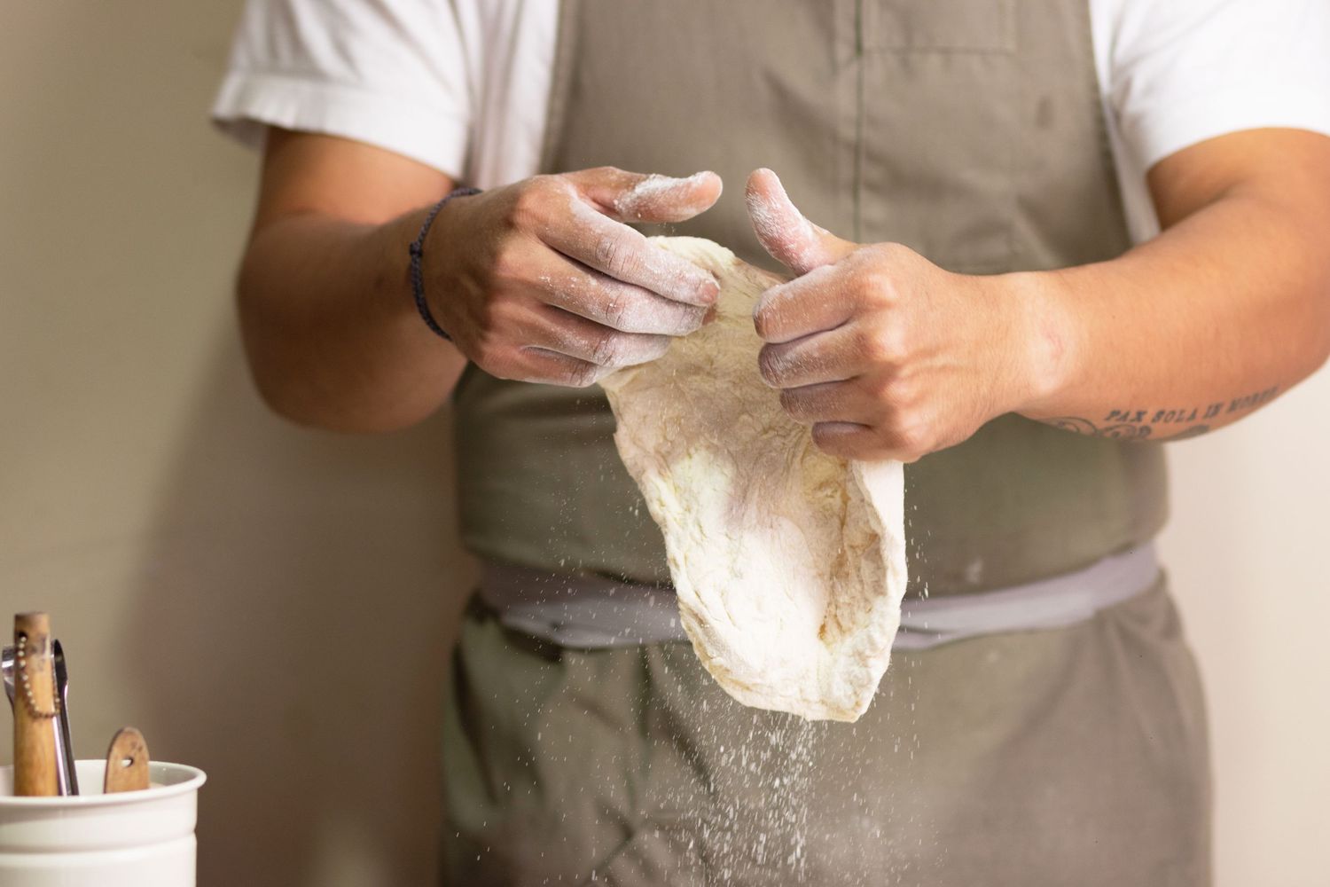 Baker in apron stretching fresh dough with flour-dusted hands in a professional kitchen setting.
