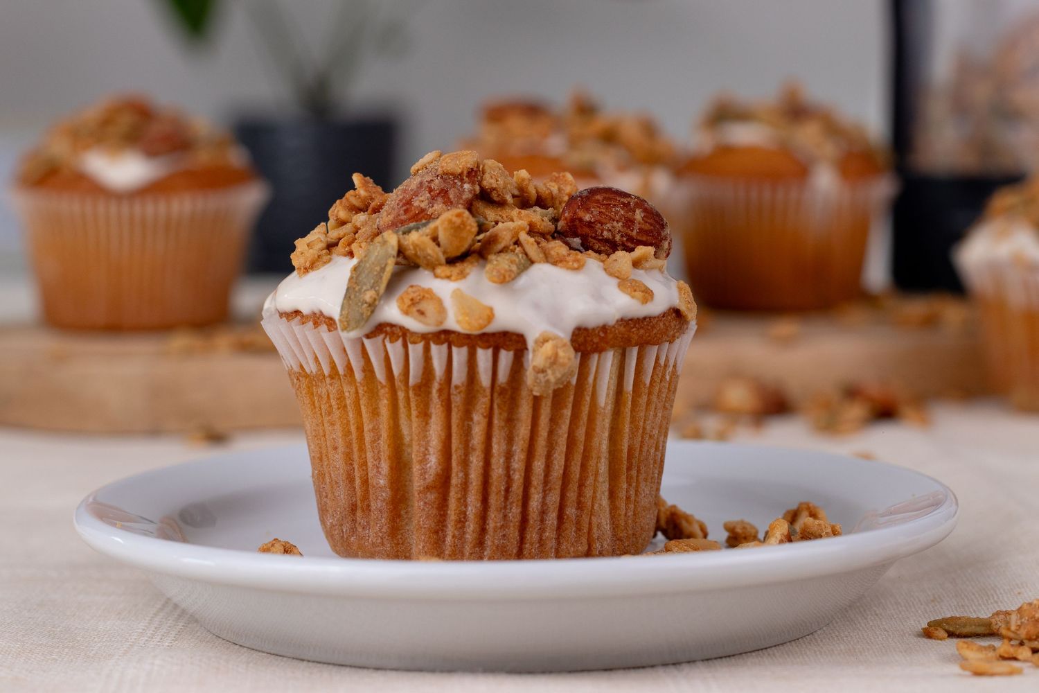 A frosted carrot muffin topped with granola and pecans on a white plate, with more muffins in the background.