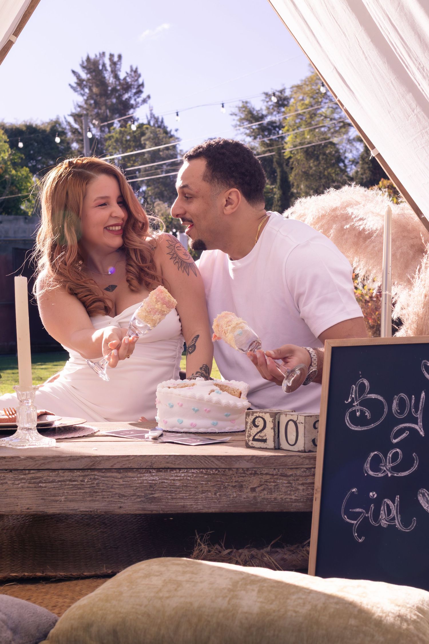 Couple laughing and sharing cake at a gender reveal party with a Boy or Girl chalkboard sign outdoors.
