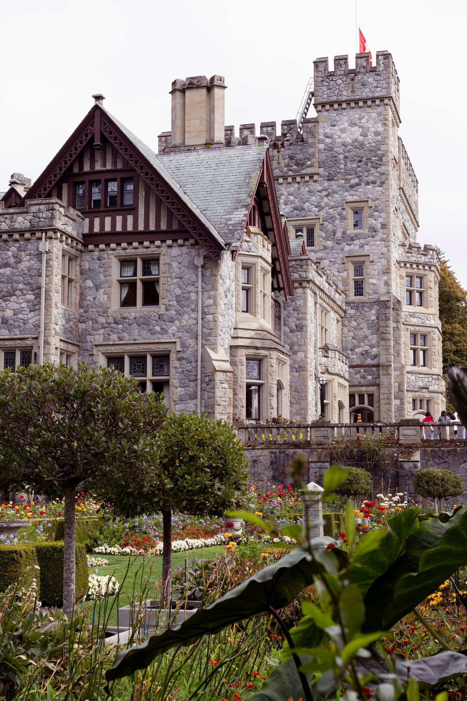 Gothic stone castle with Tudor-style architecture features beautiful flower gardens and landscaping along its facade.