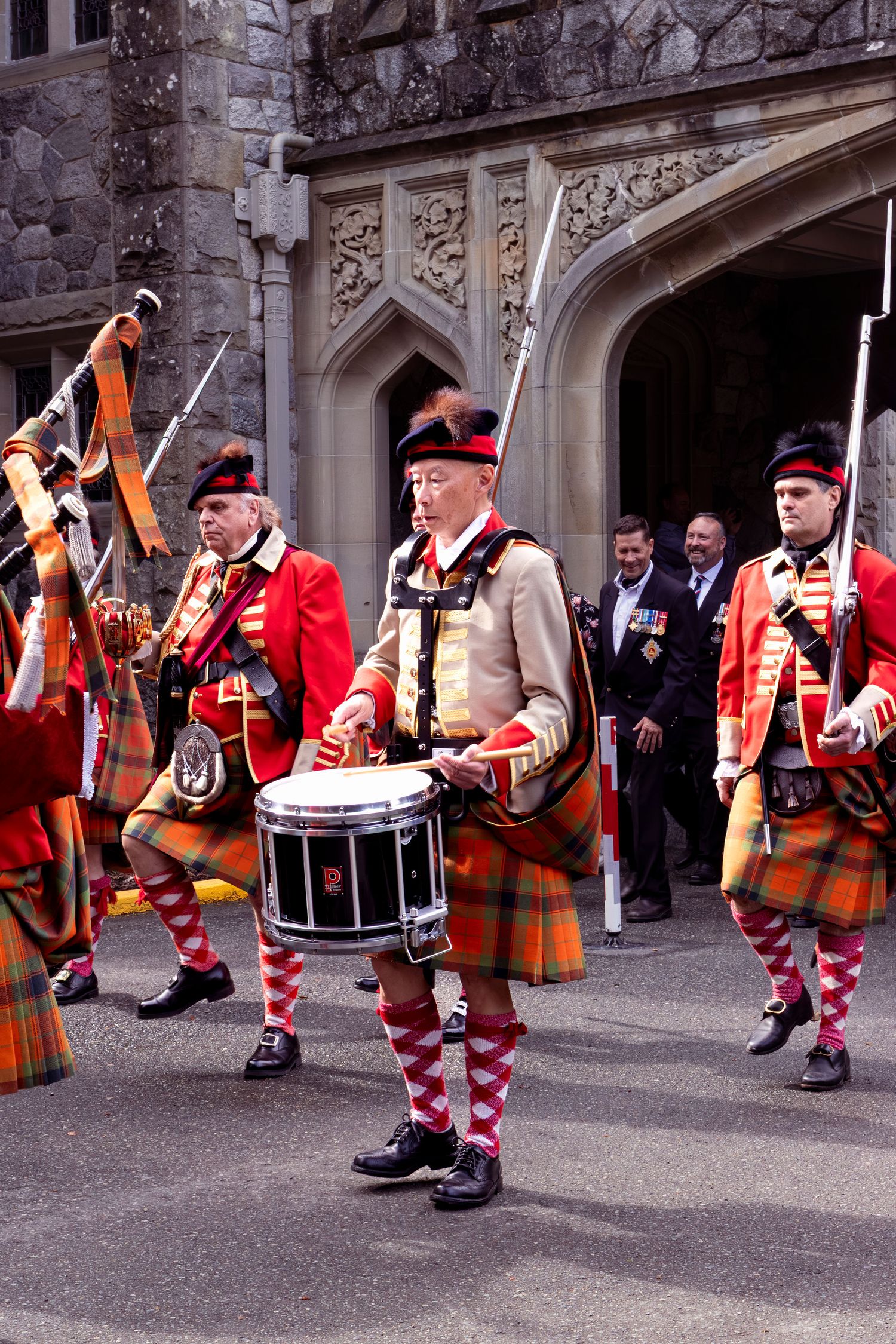 Scottish military band performers in traditional kilts and red uniforms with drums and bagpipes at historic stone archway.