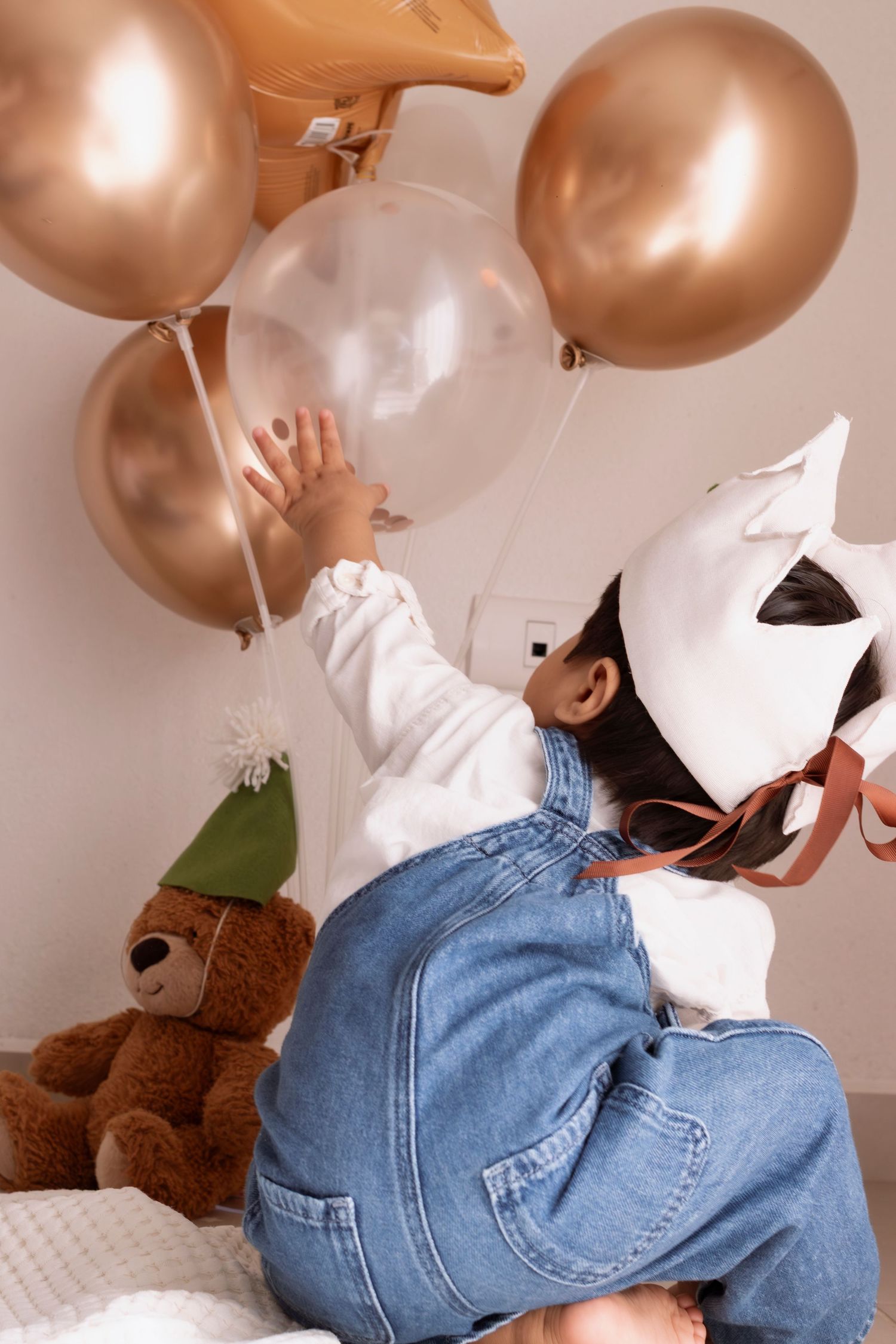Baby in denim overalls crawls on floor while reaching for gold and white balloons in a cozy room.