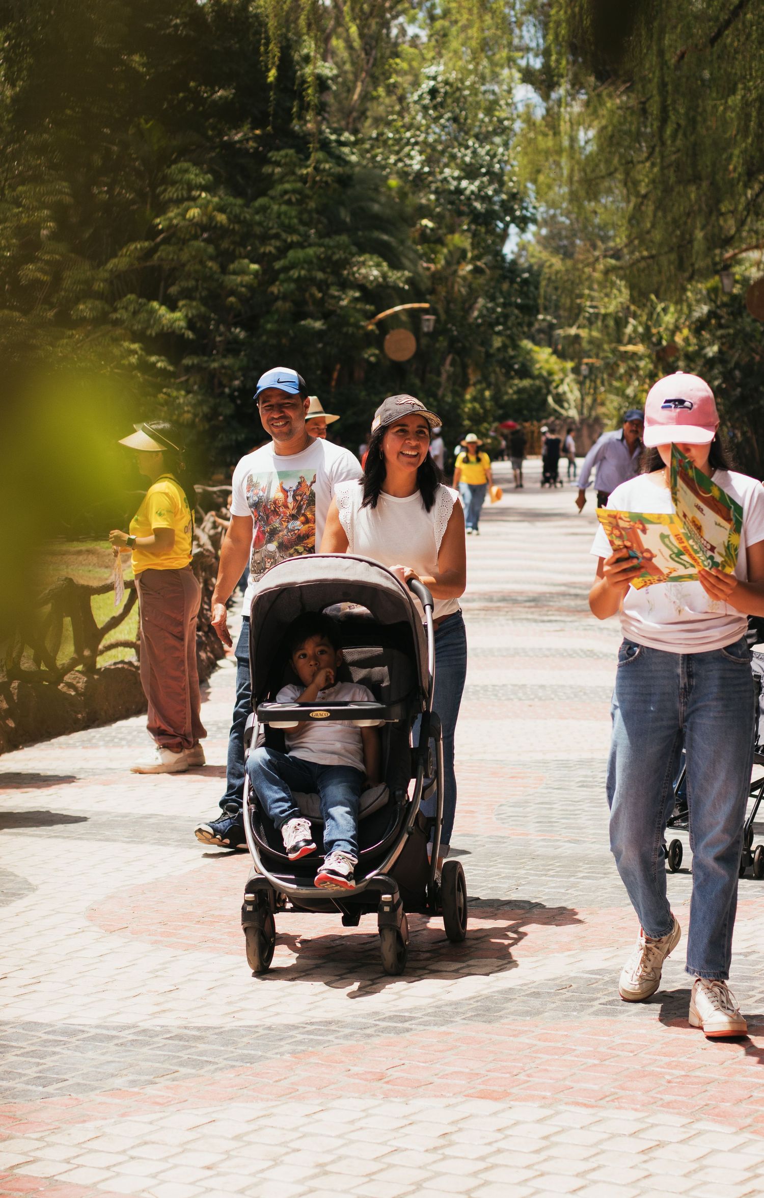 Family strolling on a sunny park path, parents pushing a baby stroller with a child seated inside.