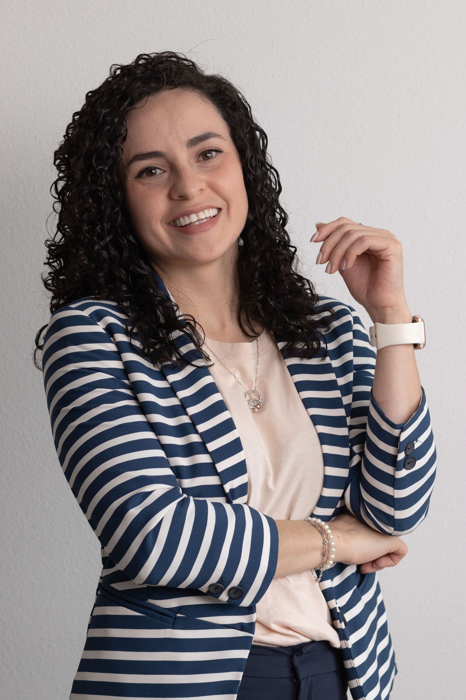Smiling woman with curly hair wearing a blue striped blazer over a pink top, posing confidently against a white wall.