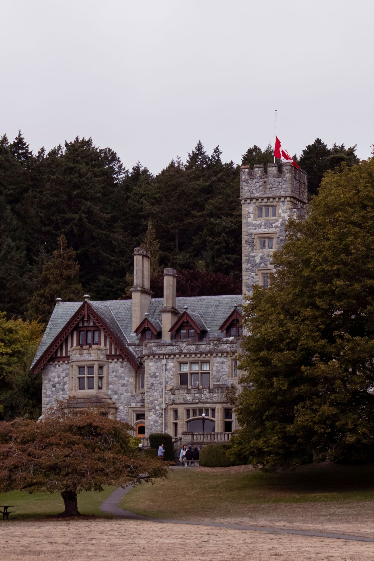 Gothic stone castle with turrets and gabled windows stands among autumn trees in a scenic woodland setting.