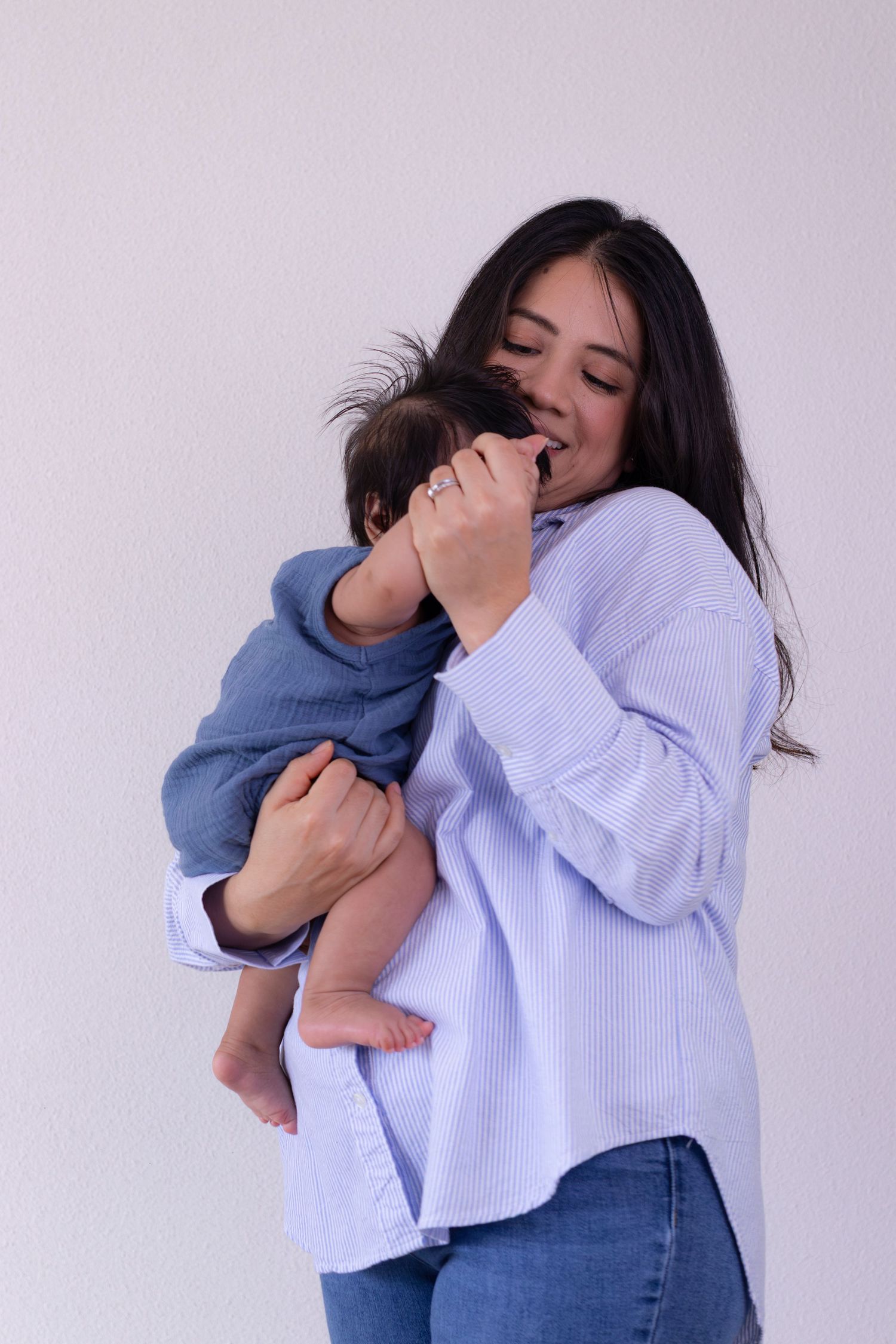 Young mother in blue striped shirt tenderly holding and kissing her newborn baby against white background.