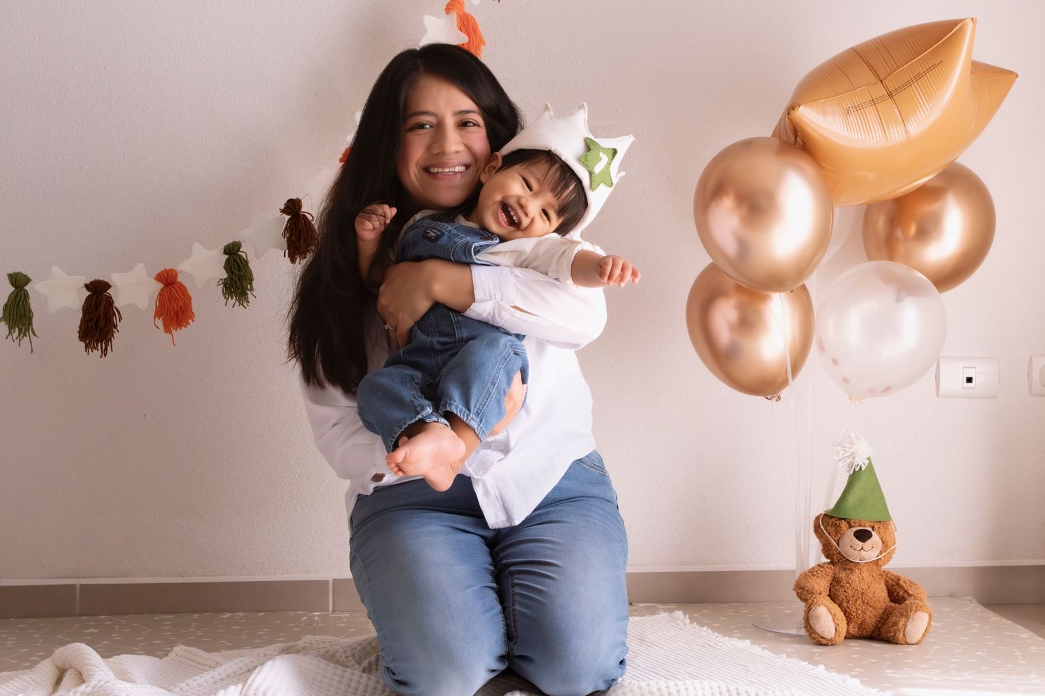 Mother hugging smiling baby in dinosaur hat at first birthday party with gold balloons and teddy bear.