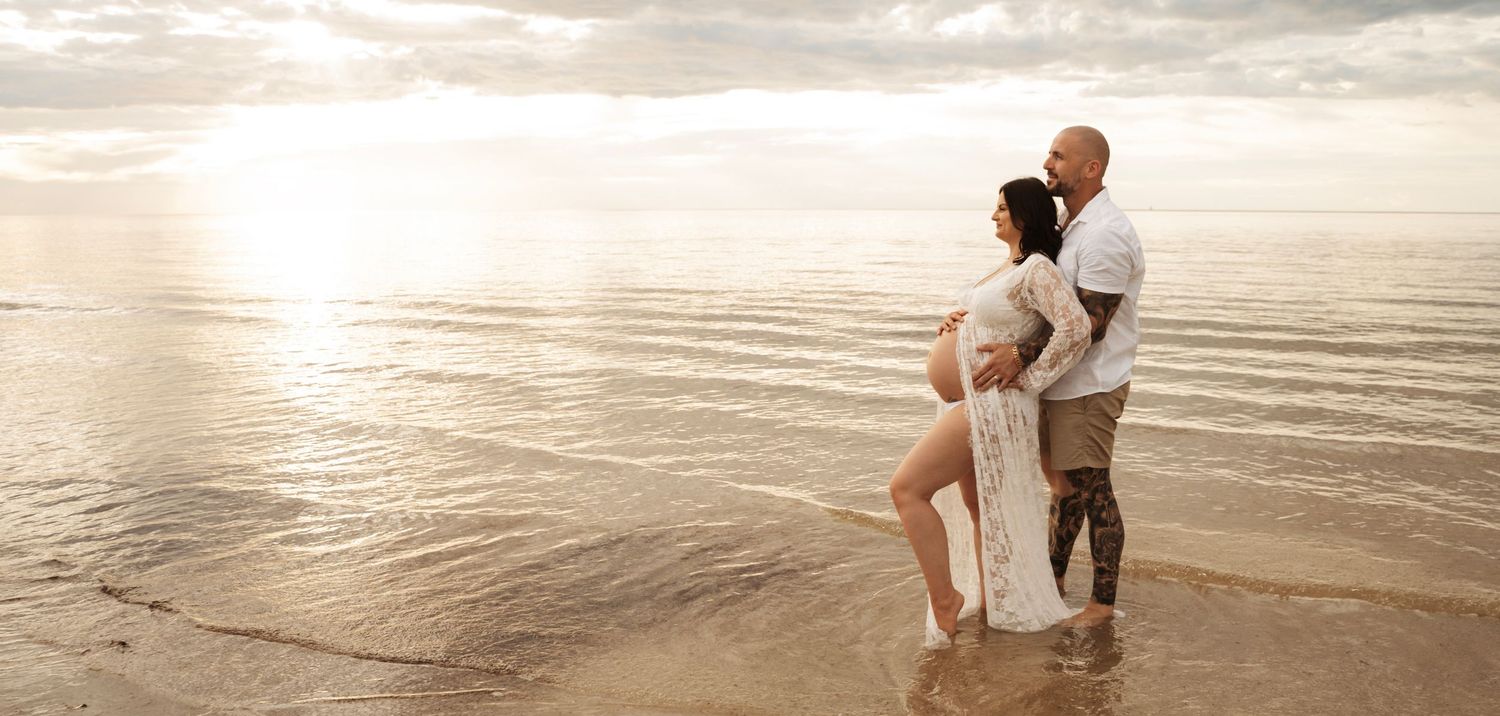 Pregnant couple standing on a serene beach at sunset, woman in white dress, man with tattoos, waves gently lapping their feet.