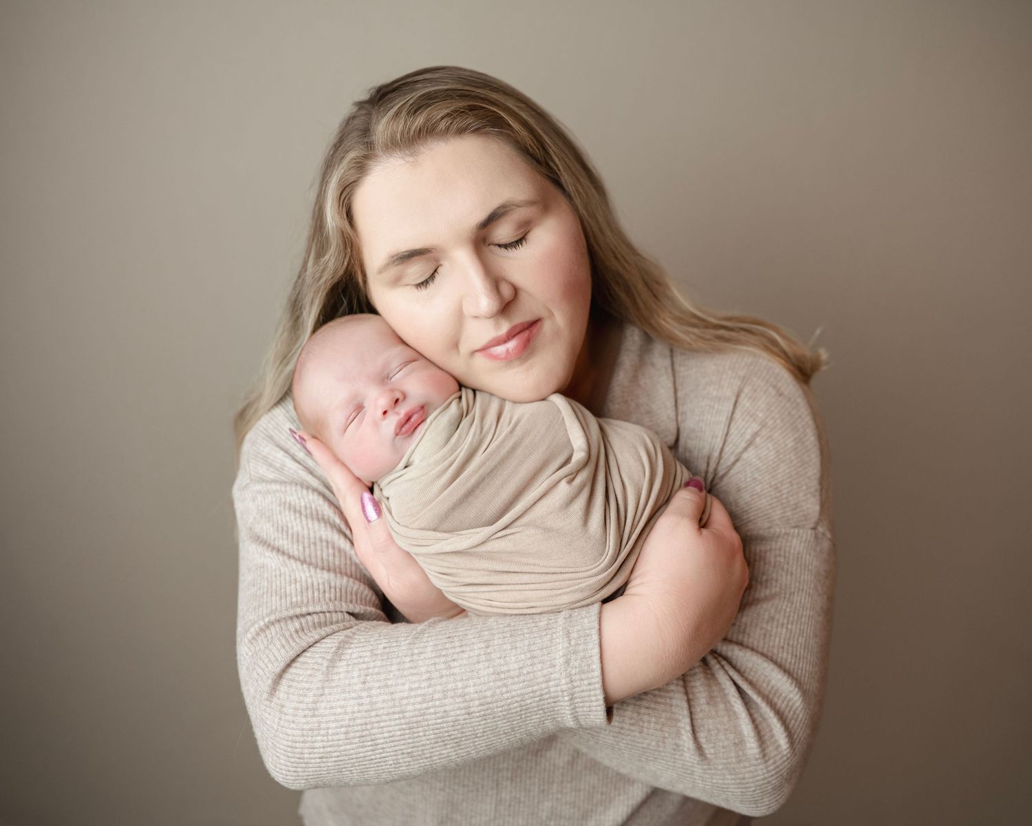 Mother tenderly holds sleeping newborn baby wrapped in beige swaddle against her chest in a studio portrait.