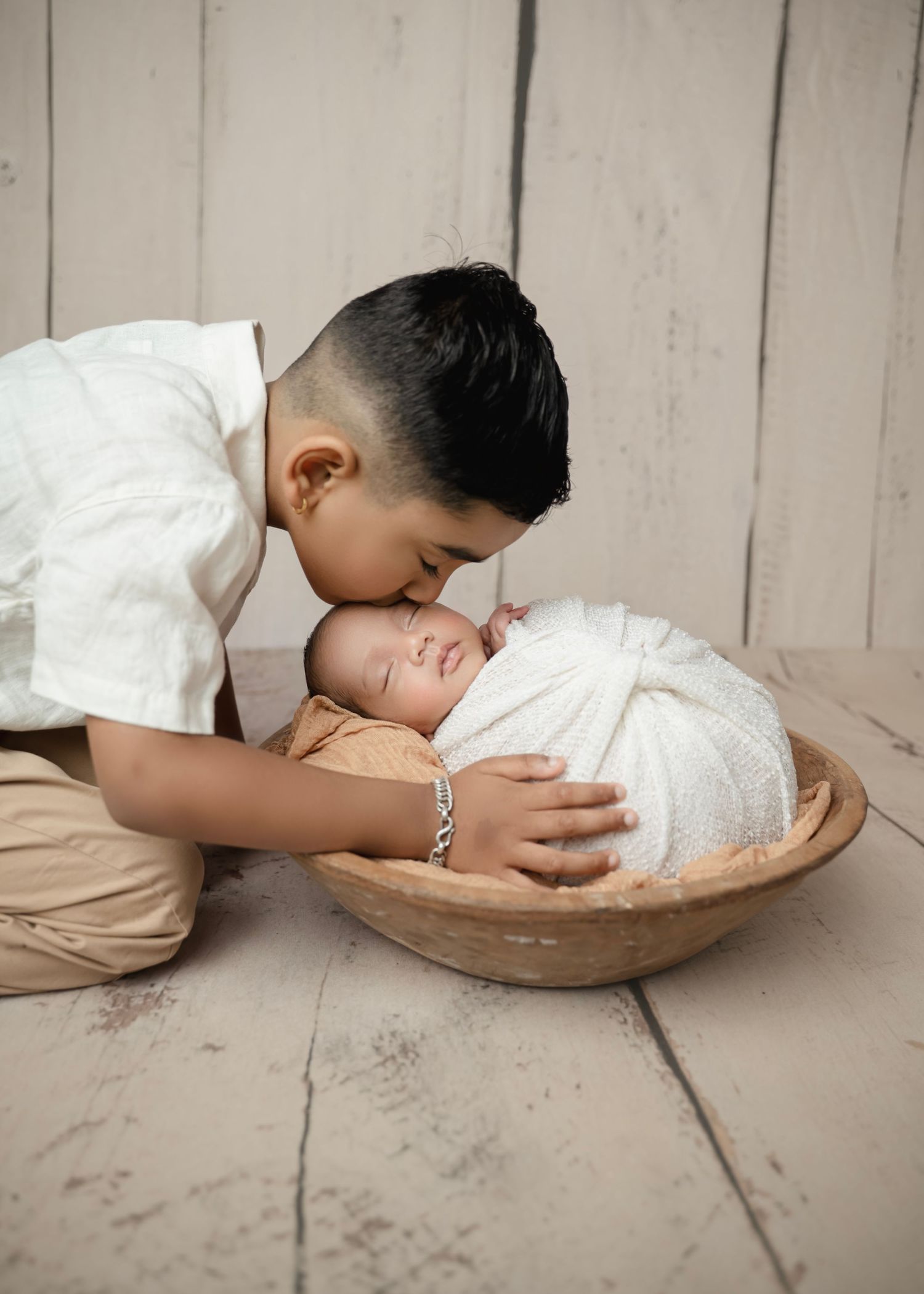 Older boy gently kissing sleeping newborn baby wrapped in white swaddle inside rustic wooden bowl.