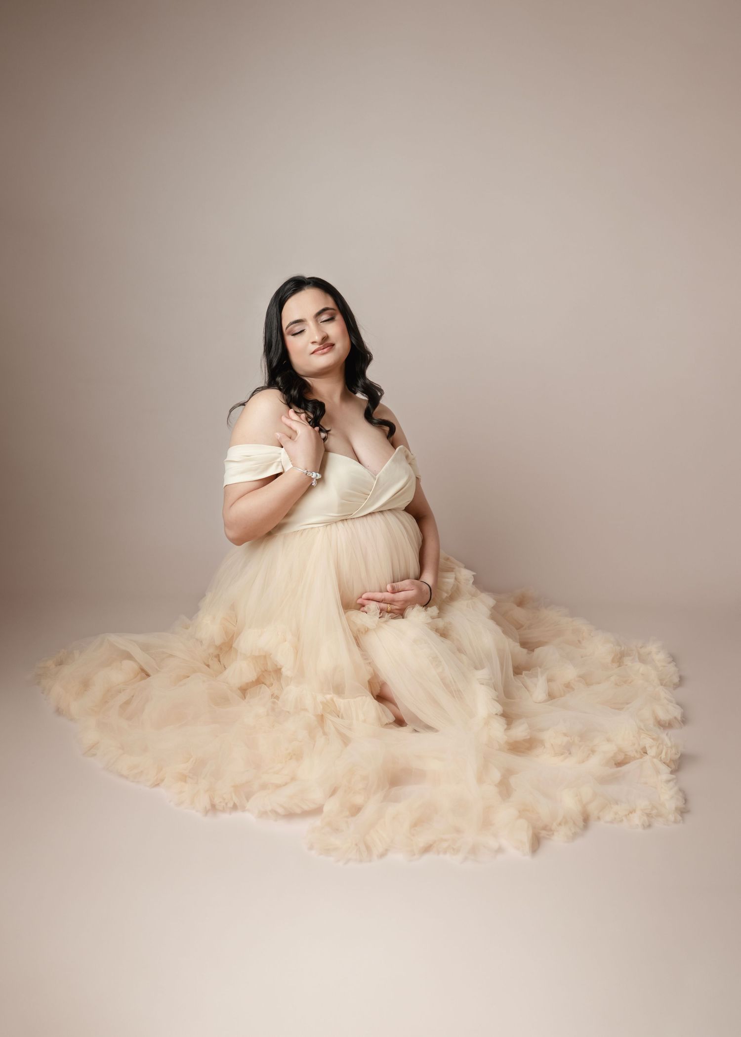 Pregnant woman in elegant cream tulle off-shoulder gown sitting gracefully on studio floor during maternity photoshoot.