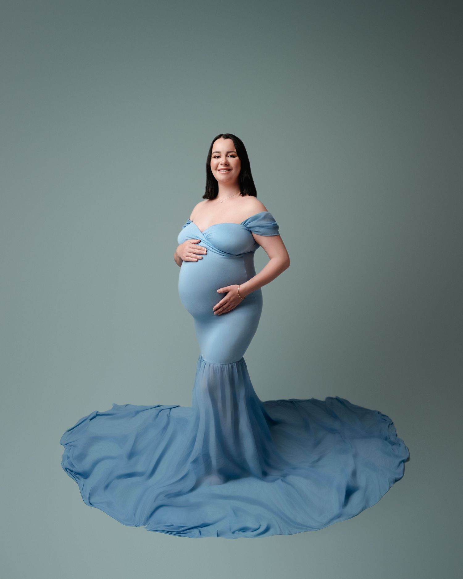 Pregnant woman in elegant blue off-shoulder mermaid gown with flowing train poses for maternity studio portrait.