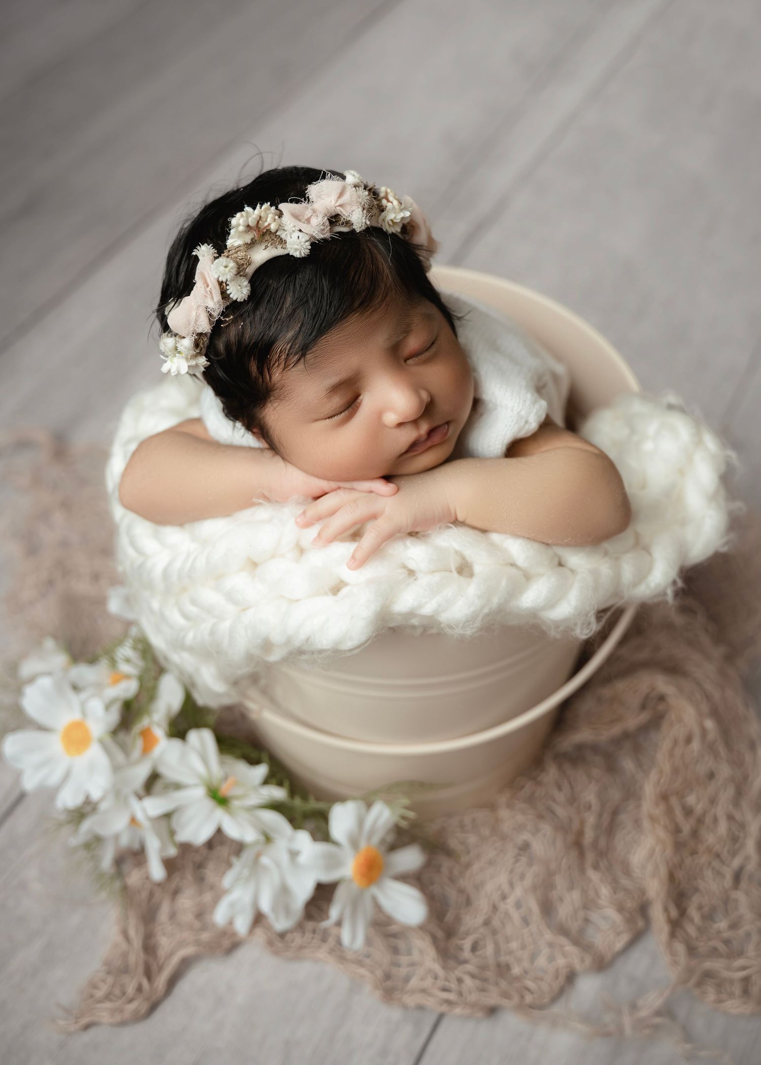Sleeping newborn baby in a cream bucket with white fluffy blanket, floral crown, and daisy flowers on a wooden floor.