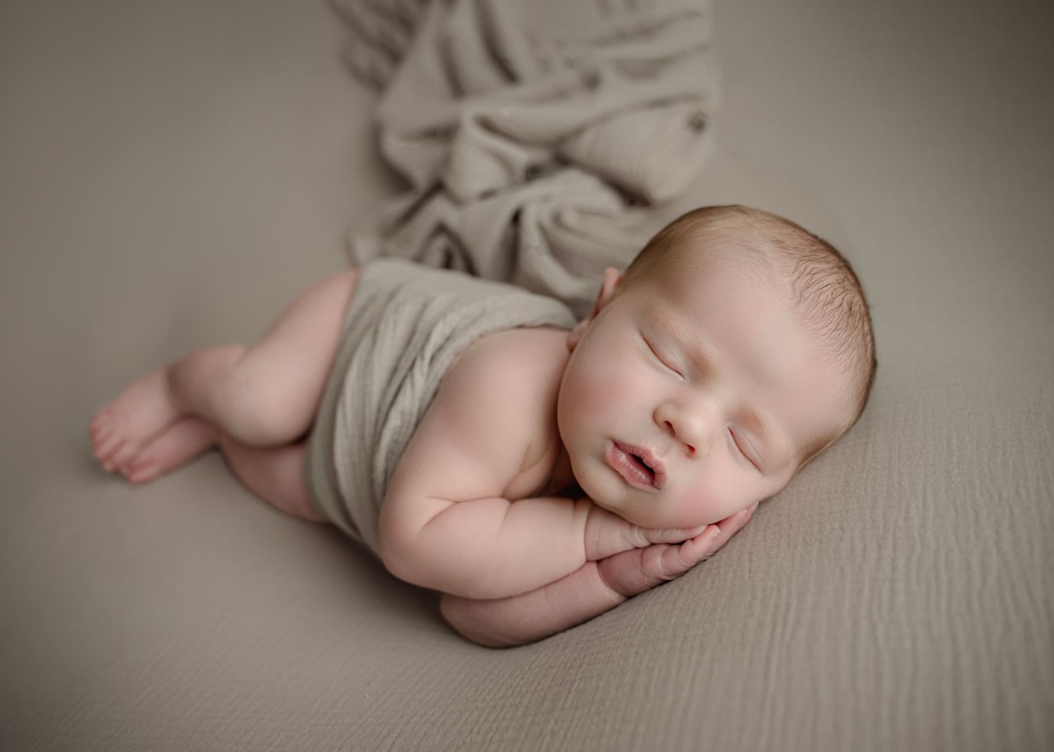 Peaceful newborn baby sleeping curled up on a soft beige blanket, wrapped in a light gray swaddle cloth.