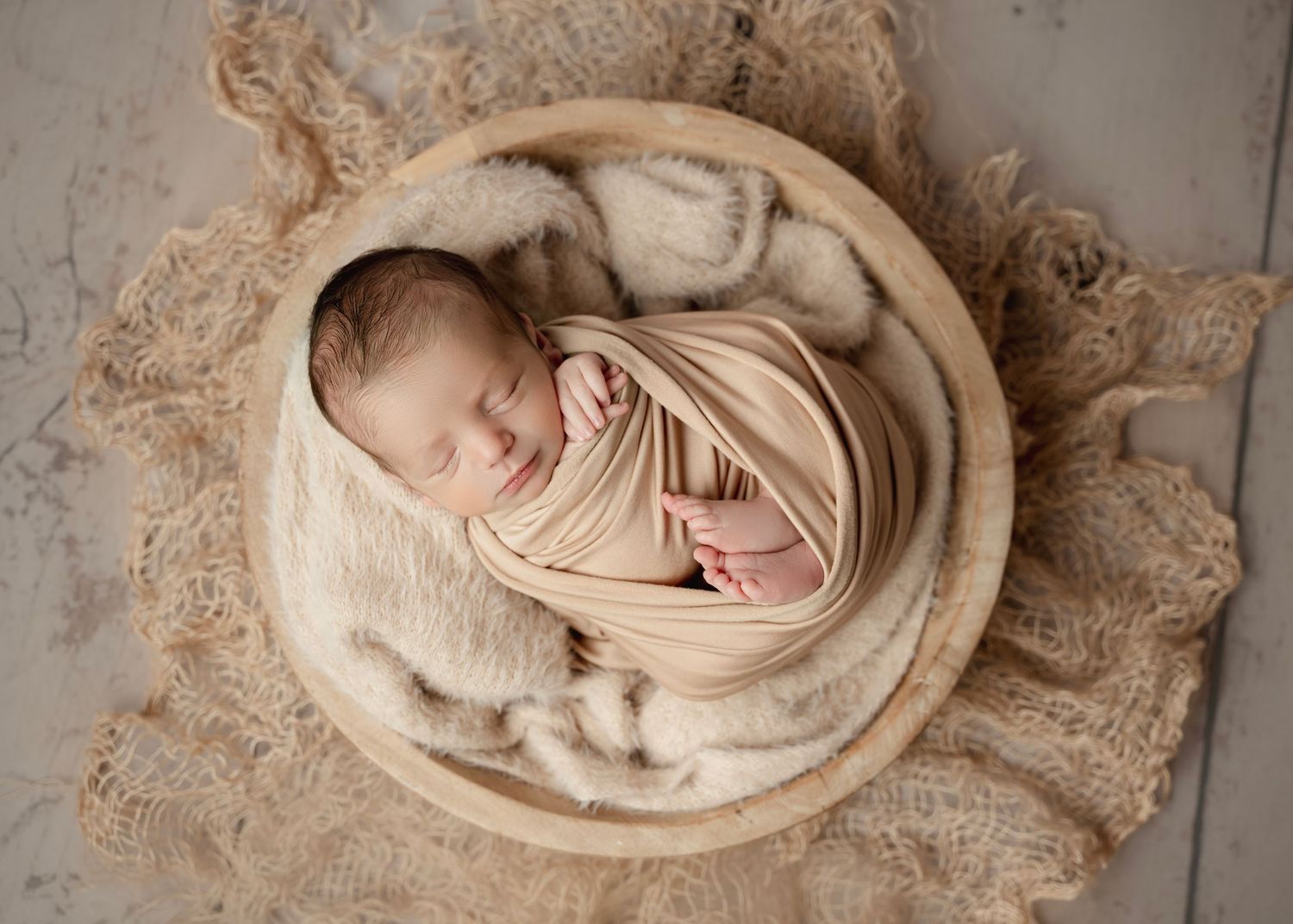 Newborn baby swaddled in beige wrap, sleeping peacefully in a wooden bowl on a lace blanket.