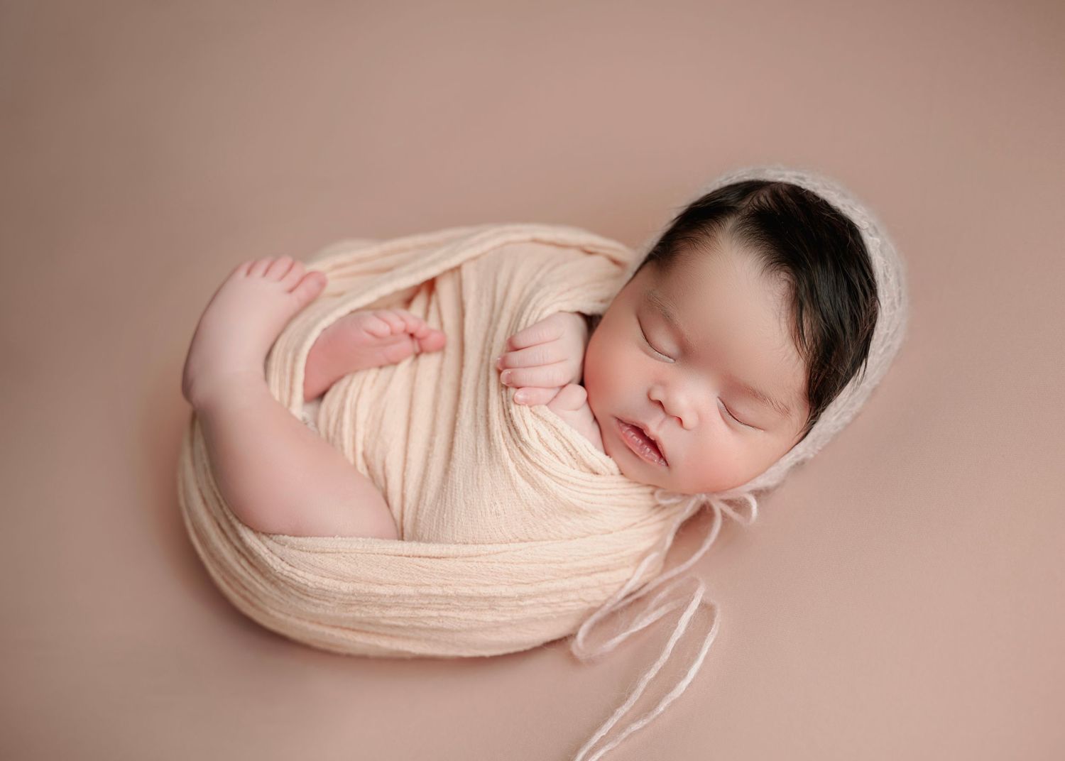 Sleeping newborn baby swaddled in cream wrap wearing a bonnet, posed on a beige background in a studio portrait.