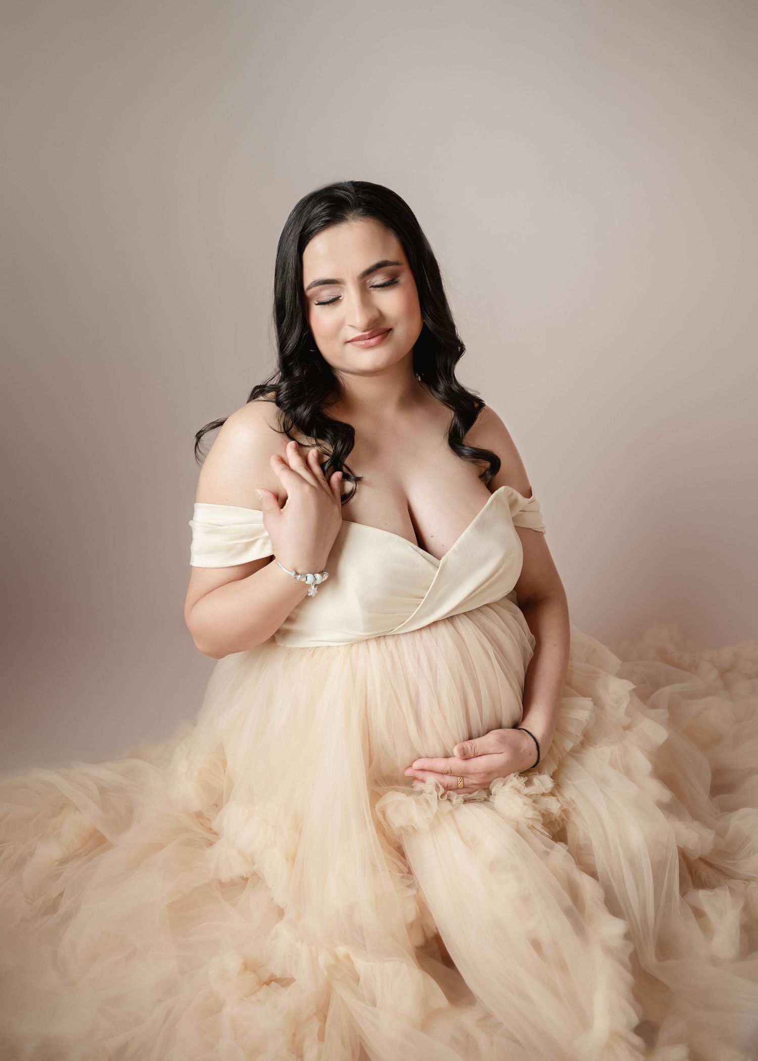 Pregnant woman in elegant off-shoulder champagne tulle gown cradling her baby bump in a soft studio maternity portrait.