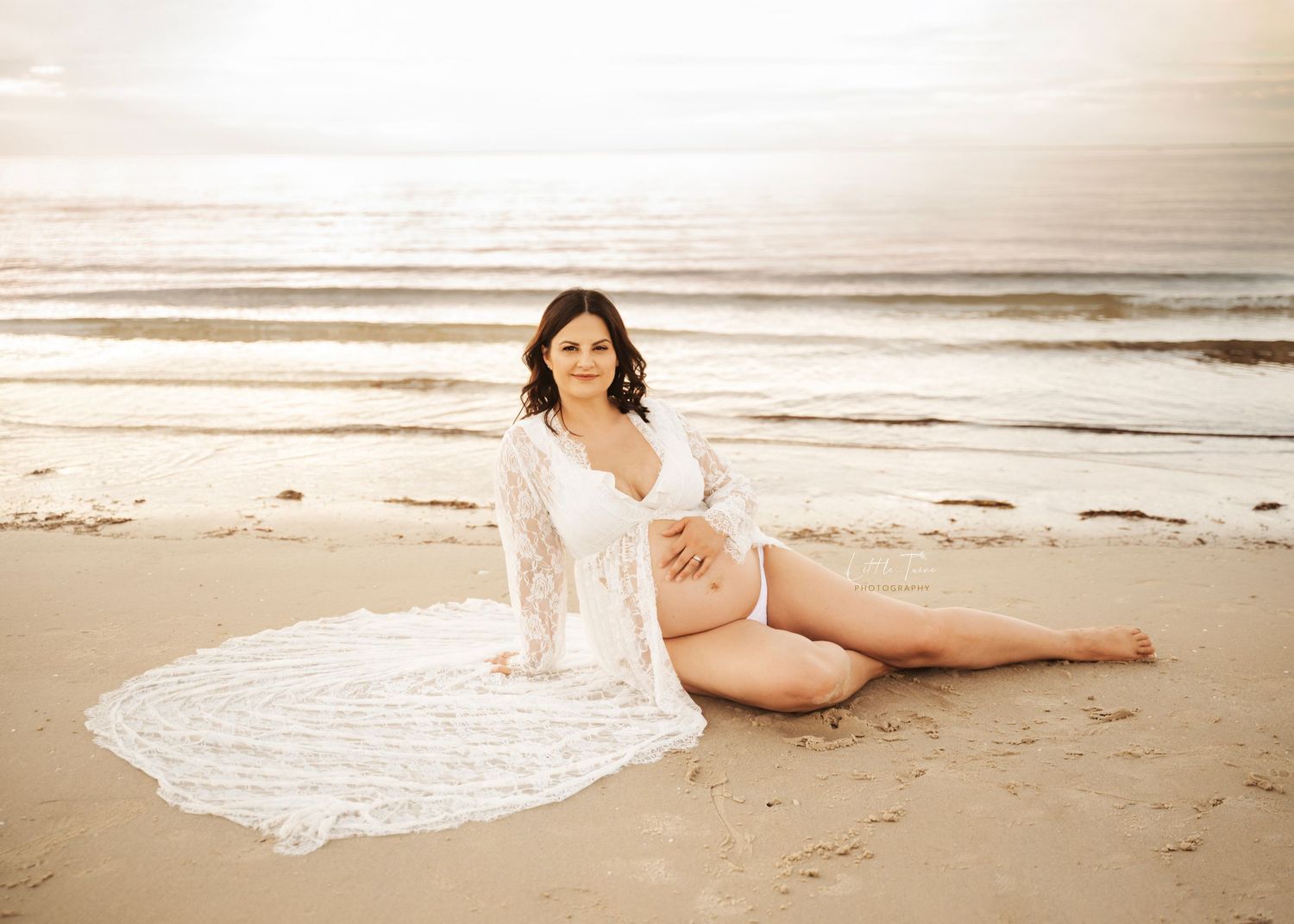 Pregnant woman in white lace robe sits on sandy beach at sunset, cradling baby bump in a serene maternity photo.