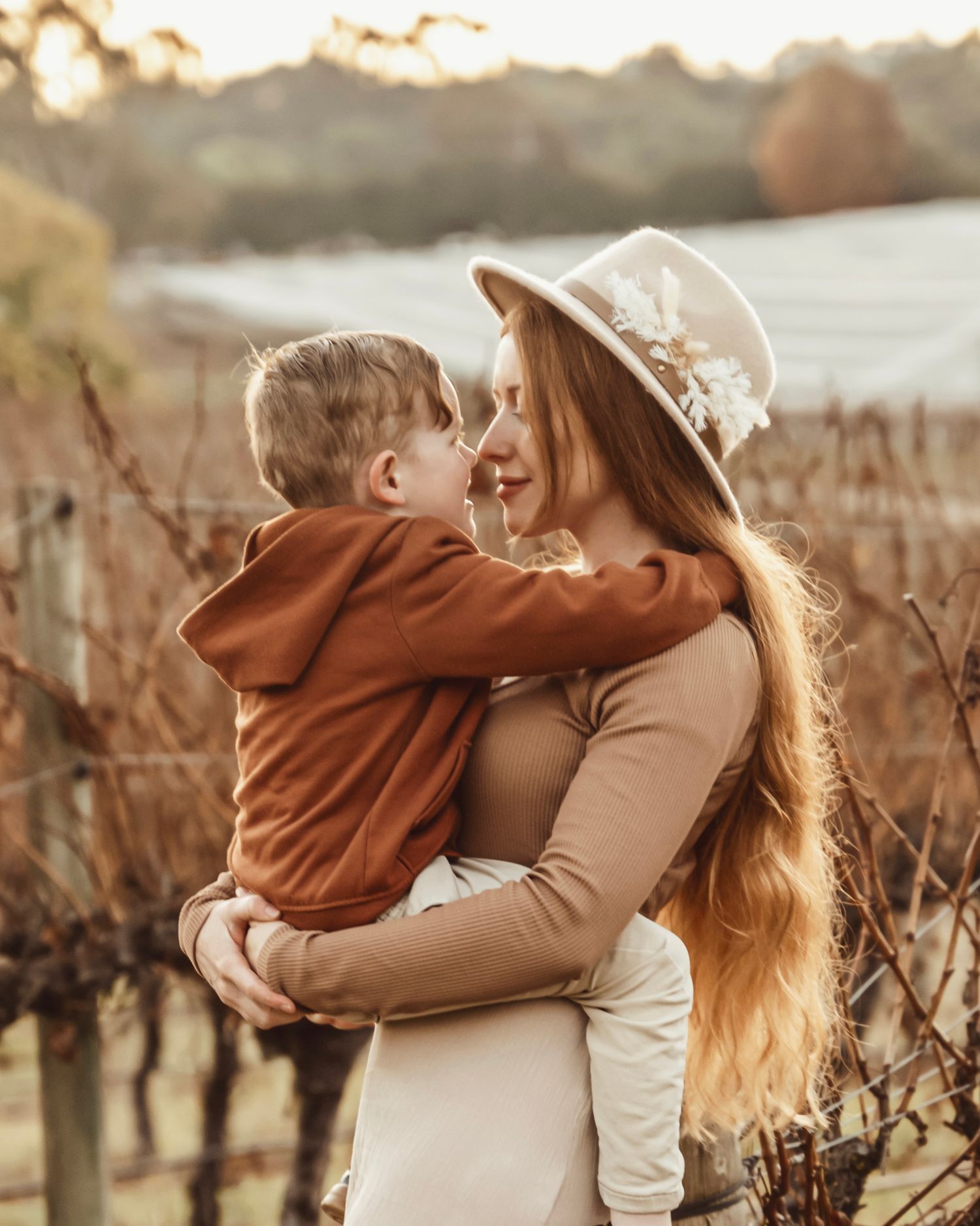 A mother with long red hair and a floral hat holds her young son in a vineyard during golden hour.