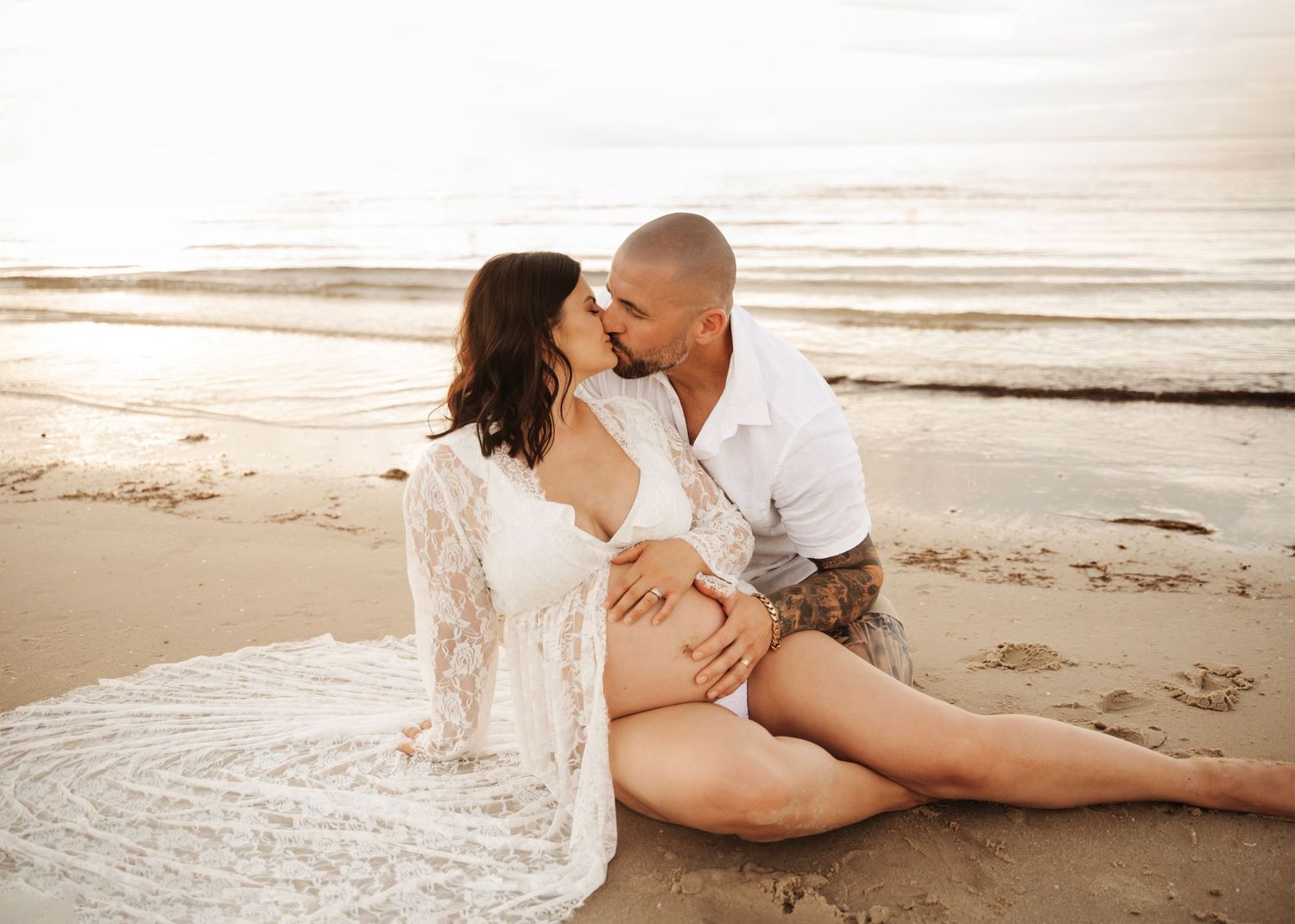 Couple sharing a tender kiss on the beach during a golden sunset maternity photoshoot by the ocean.