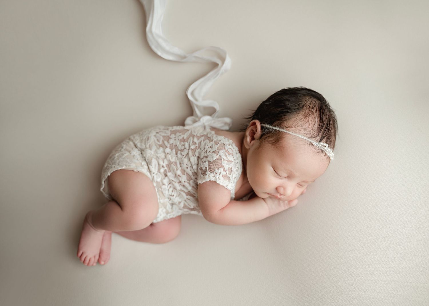 Sleeping newborn baby in white lace romper and headband posed on neutral background.
