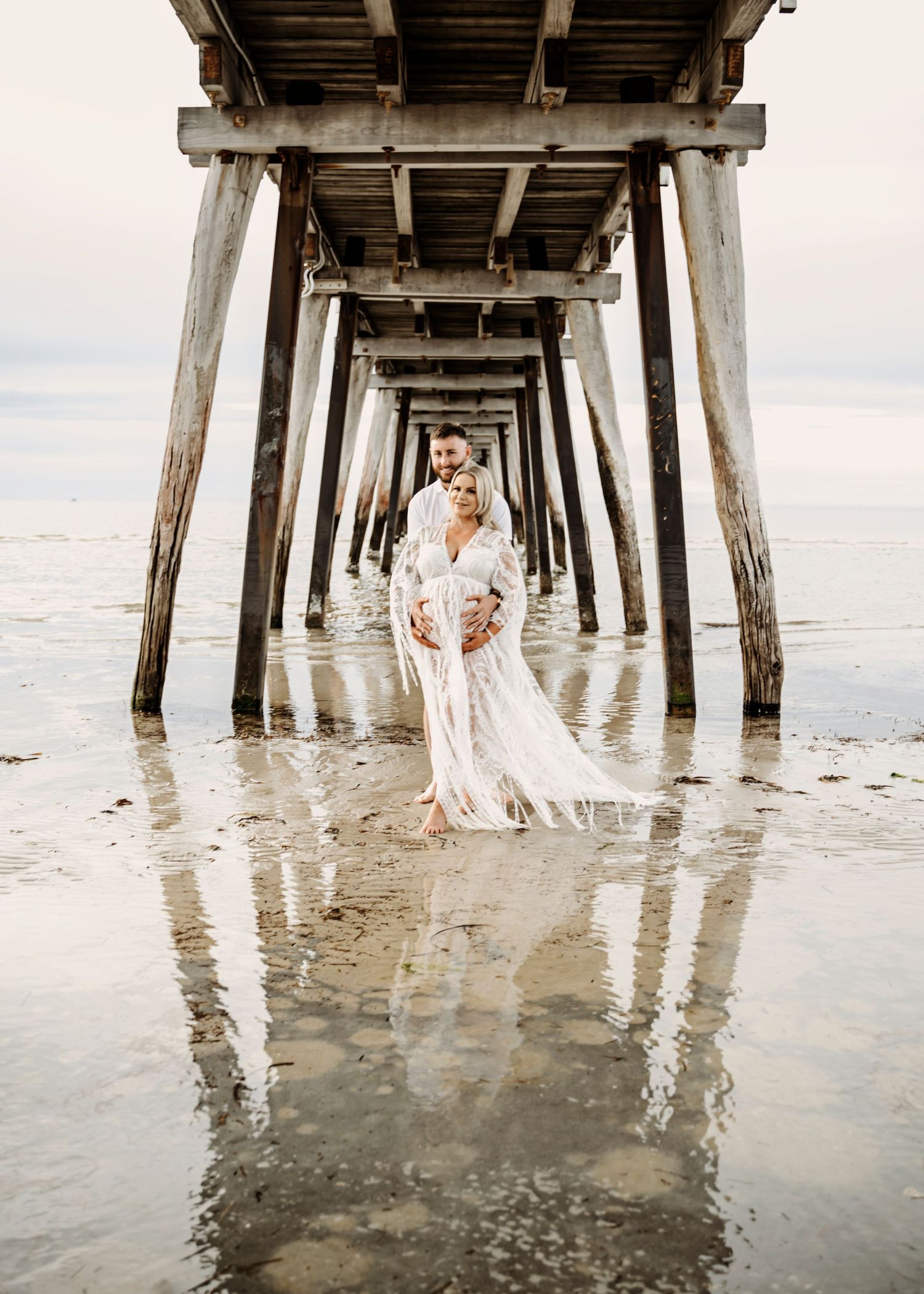 Couple embracing under a wooden pier on wet sand, woman in flowing white lace gown, romantic beach maternity photo.