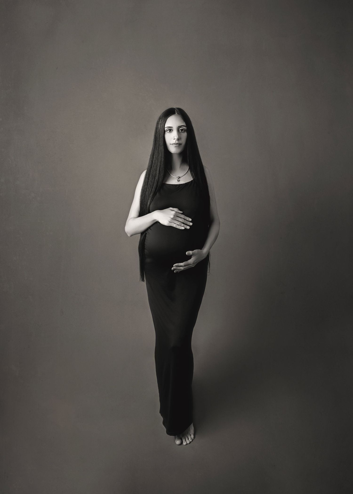 Black and white maternity photo of a woman in a black dress with long dark hair cradling her baby bump in a studio.